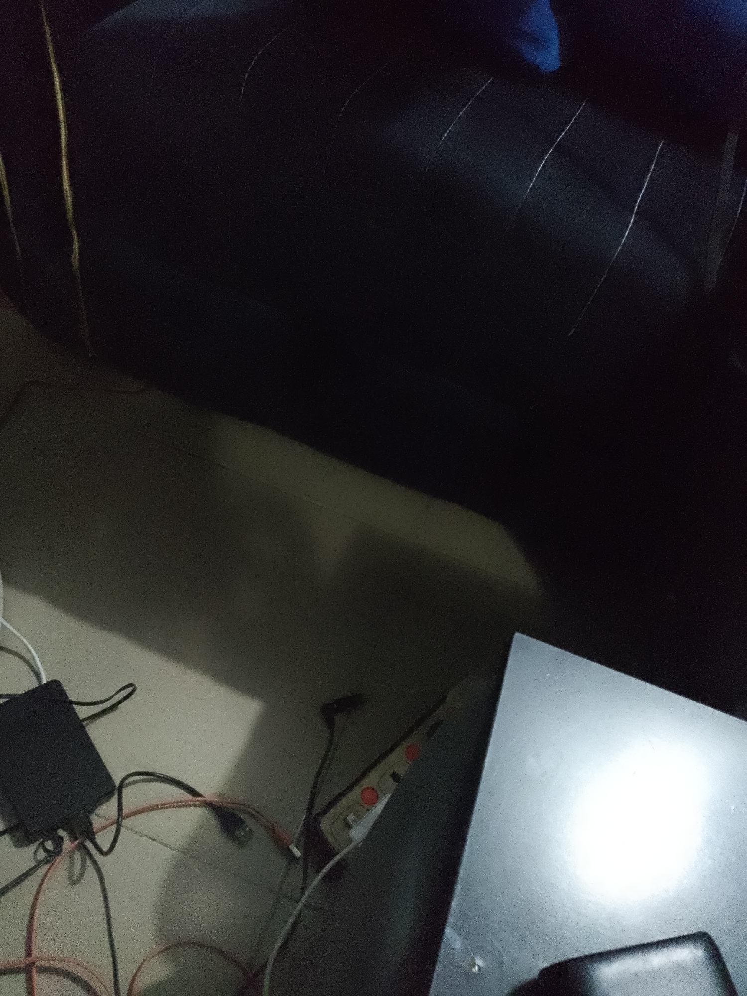 An overhead shot shows a dark, tufted sofa on the right side of the image, with electrical cords and power strips on the floor in front of it. A black power brick is visible, with multiple cords plugged into it and others extending from it. A power strip with a red button is also on the floor, with a cord plugged into it. The floor appears to be tiled or concrete. To the right of the cords and power strip, a dark, angled surface with a bright light reflection is visible, possibly a table or desk. The overall lighting is dim.