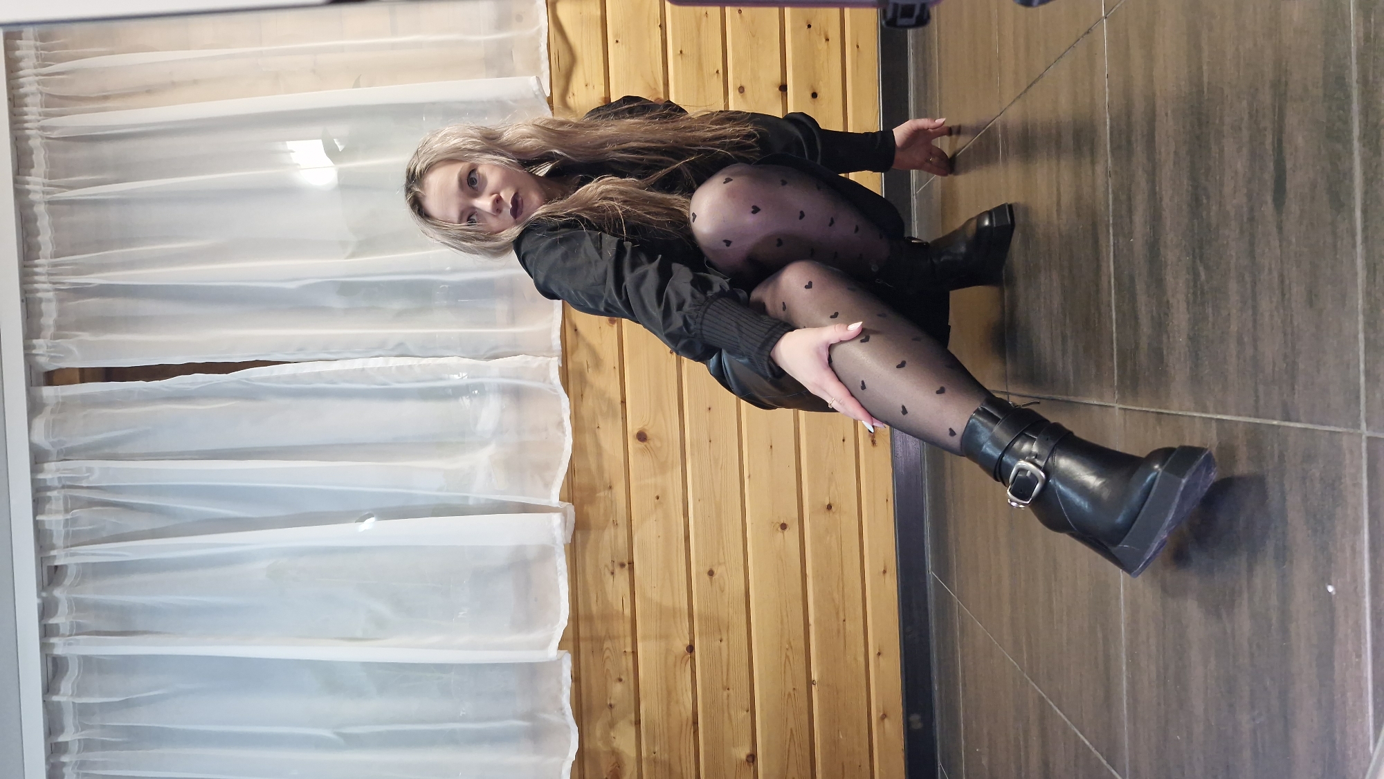 This image captures a young woman seated on an indoor floor, likely in Otchor, Russia. She is positioned against a light-toned wooden panel wall, with a sheer white curtain partially visible to her left, possibly indicating a window or room divider. The floor is covered with large, dark-colored tiles.
The woman has long, straight blonde or light brown hair and gazes directly at the camera with a neutral to slightly serious expression. She wears dark lipstick. Her attire consists of a black long-sleeved top or jacket with ribbed cuffs, and sheer black pantyhose or tights patterned with small black hearts. She is wearing black, chunky platform boots with a prominent buckle strap around the ankle.
Her pose is a seated one; her left leg is bent at the knee, while her right leg is extended forward. Her left hand rests gently on her right thigh. The scene appears to be a posed photograph, utilizing natural light or soft artificial lighting within the room. No visible text is present in the image.