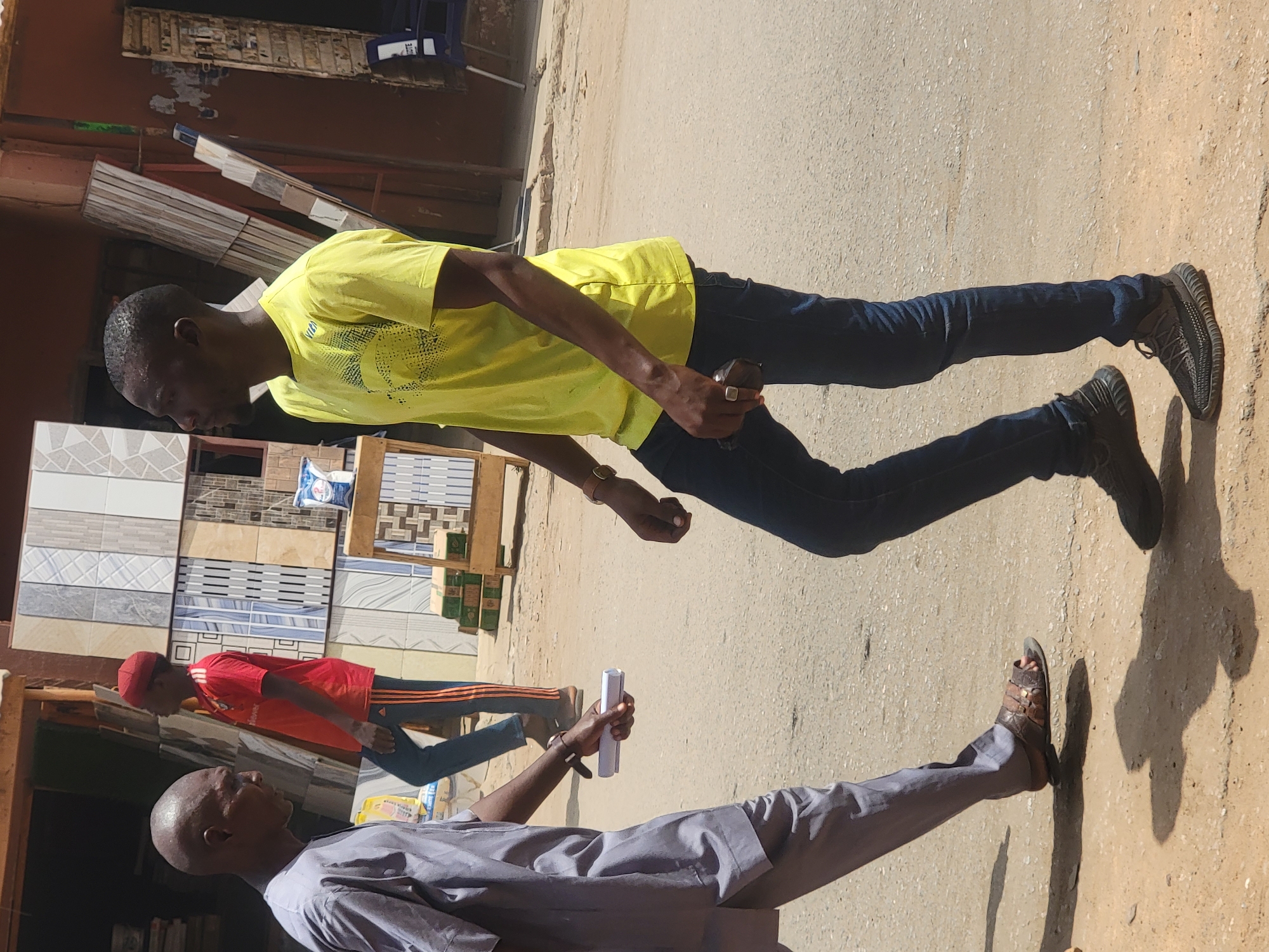 The image shows a street scene in Maiburiji, Nigeria. Three men are visible, walking on a paved surface. The man in the foreground wears a light gray outfit and sandals, carrying a rolled-up white object. Behind him, a man wears a red shirt with dark pants and a red cap. In the background, a man wears a yellow shirt and blue jeans, holding a dark object in his hand. The scene is in daylight, and various objects, including tiles and building materials, are visible in the background. The location appears to be a commercial area with shops.