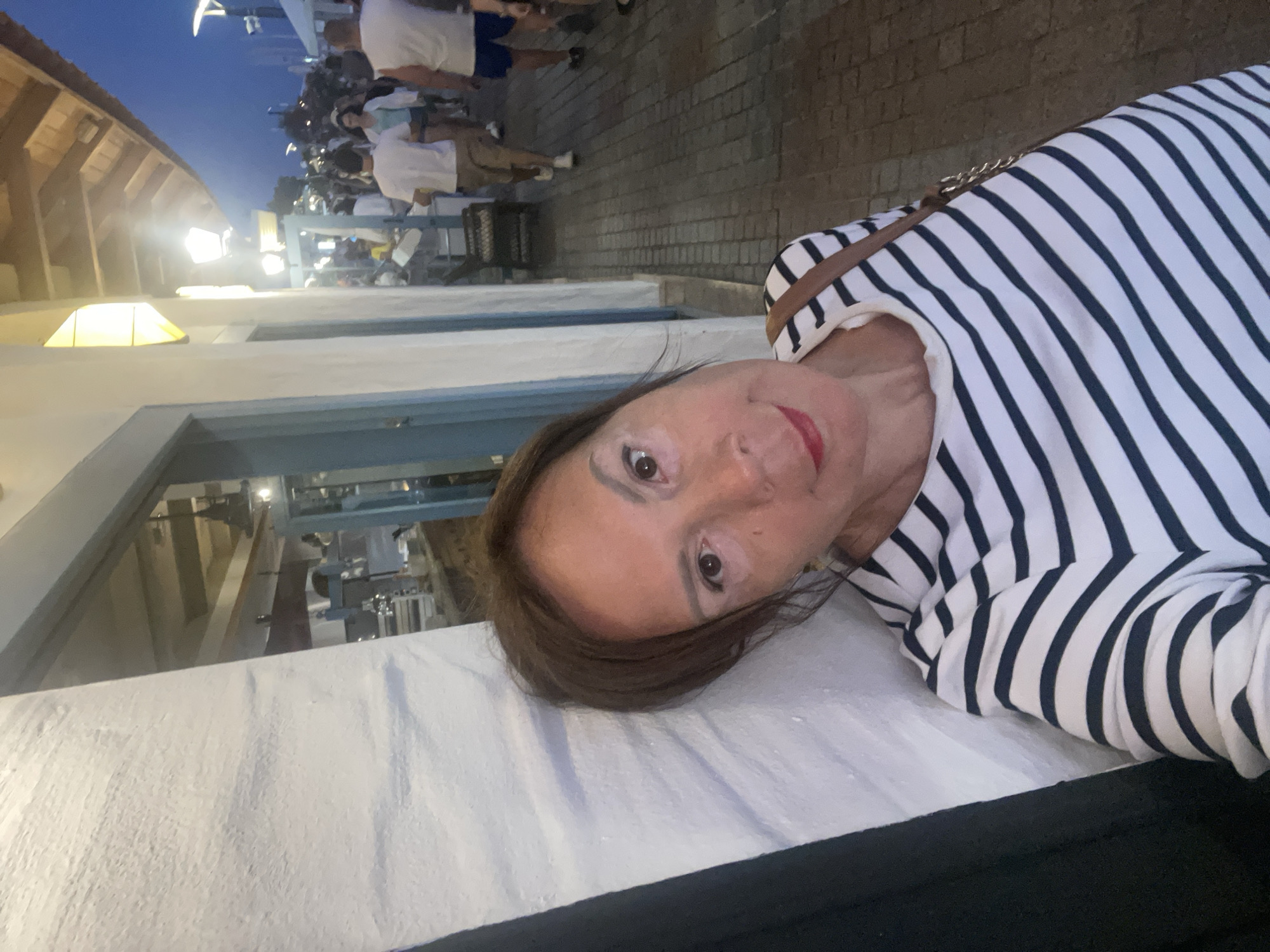 A woman is taking a selfie outdoors, leaning against a white textured wall on the left. She is wearing a black and white striped shirt and a brown purse with a chain strap is slung over her shoulder. Her head is tilted and she is smiling slightly, with red lipstick visible. Behind her, a pedestrian street is illuminated by streetlights and building lights, with several people walking in the background. The overall scene suggests an evening setting in a populated area, possibly a tourist or shopping district. The location is identified as Sochi, Russia.
