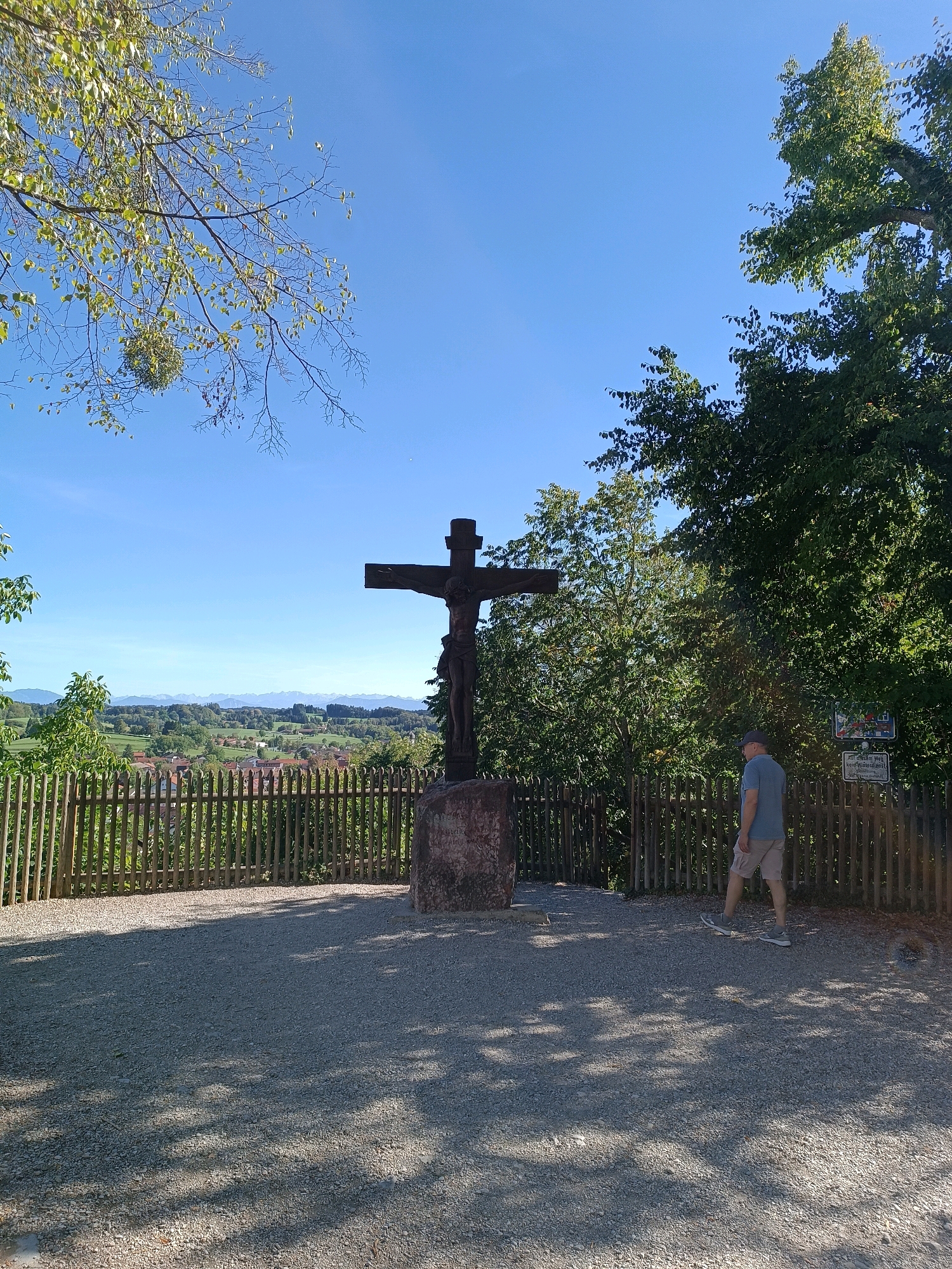 A man in a blue t-shirt and beige shorts walks on a gravel path next to a wooden fence. To the left of the man is a large wooden crucifix with a figure of Jesus on it, standing on a stone base. Behind the fence, there is a village with houses and green hills in the distance. Trees and blue sky are visible in the background. The scene appears to be in Andechs, Germany, likely on a scenic overlook.