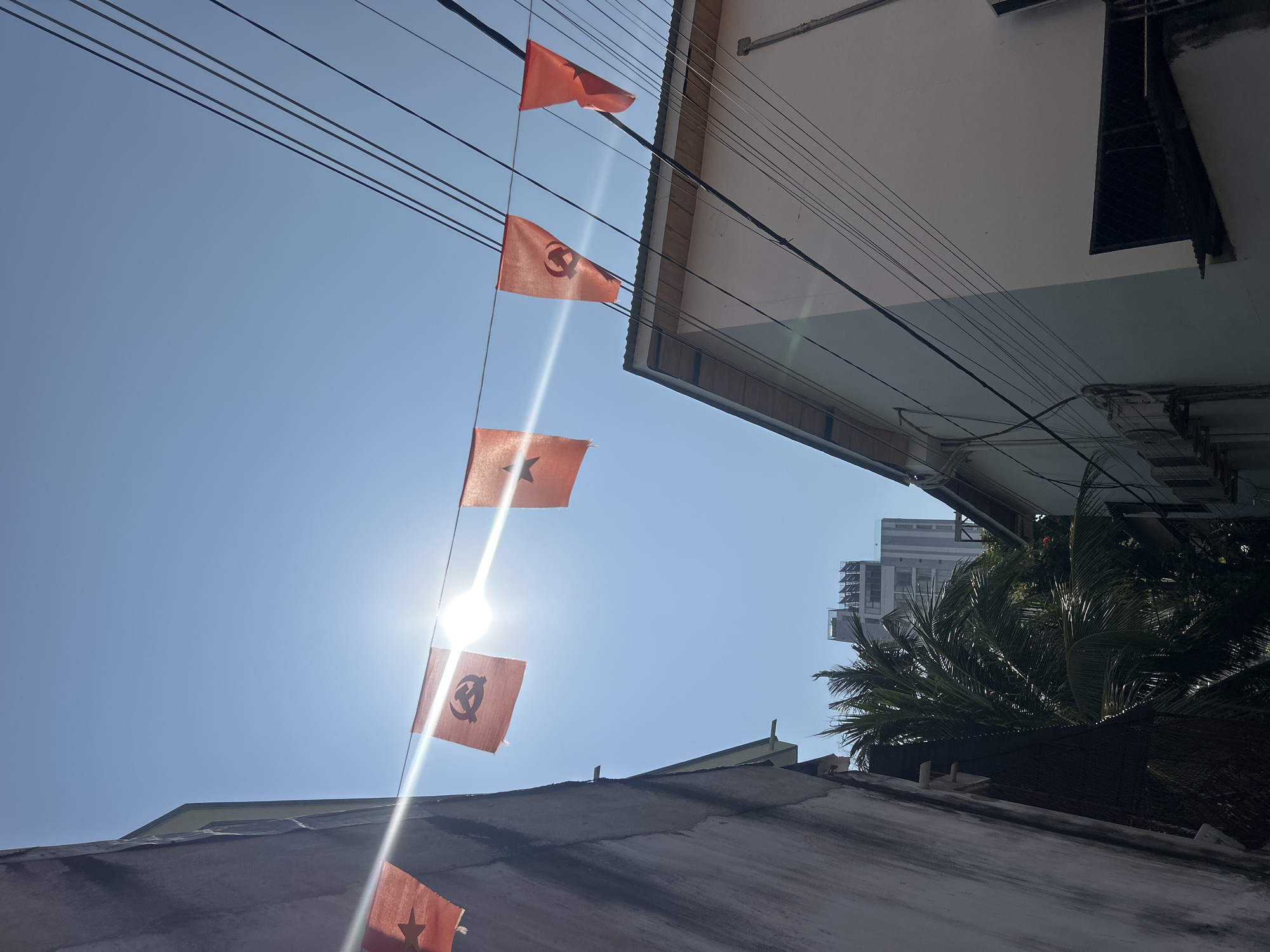 The image, taken in Bắc Nha Trang, Vietnam, shows a low-angle view of orange flags strung across power lines. The flags have symbols on them, including a star and a hammer and sickle. The sun is visible behind the flags, causing a lens flare effect. Buildings surround the area, and a palm tree is also visible.