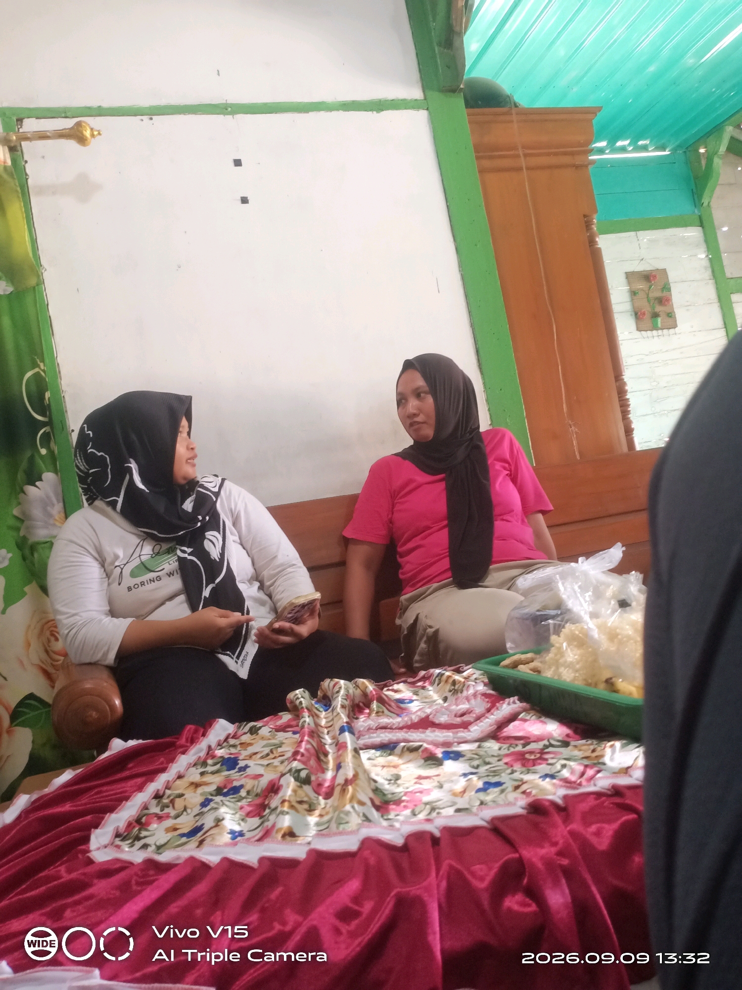 Two women are seated indoors, likely in Plosorejo, Indonesia, based on the watermark. The woman on the left wears a black hijab and a white shirt with a floral pattern and "BORING WI" visible. She holds a mobile phone. The woman on the right wears a black hijab and a bright pink t-shirt, with tan pants. They are sitting on a wooden bench. In front of them, a table is covered with a vibrant pink and floral patterned cloth. On the table, there is a green plastic container filled with snacks, possibly crackers, and a plastic bag. The room has white walls, green trim, and a turquoise corrugated metal ceiling. A wooden cabinet is visible in the background, along with a small decorative item on the wall.