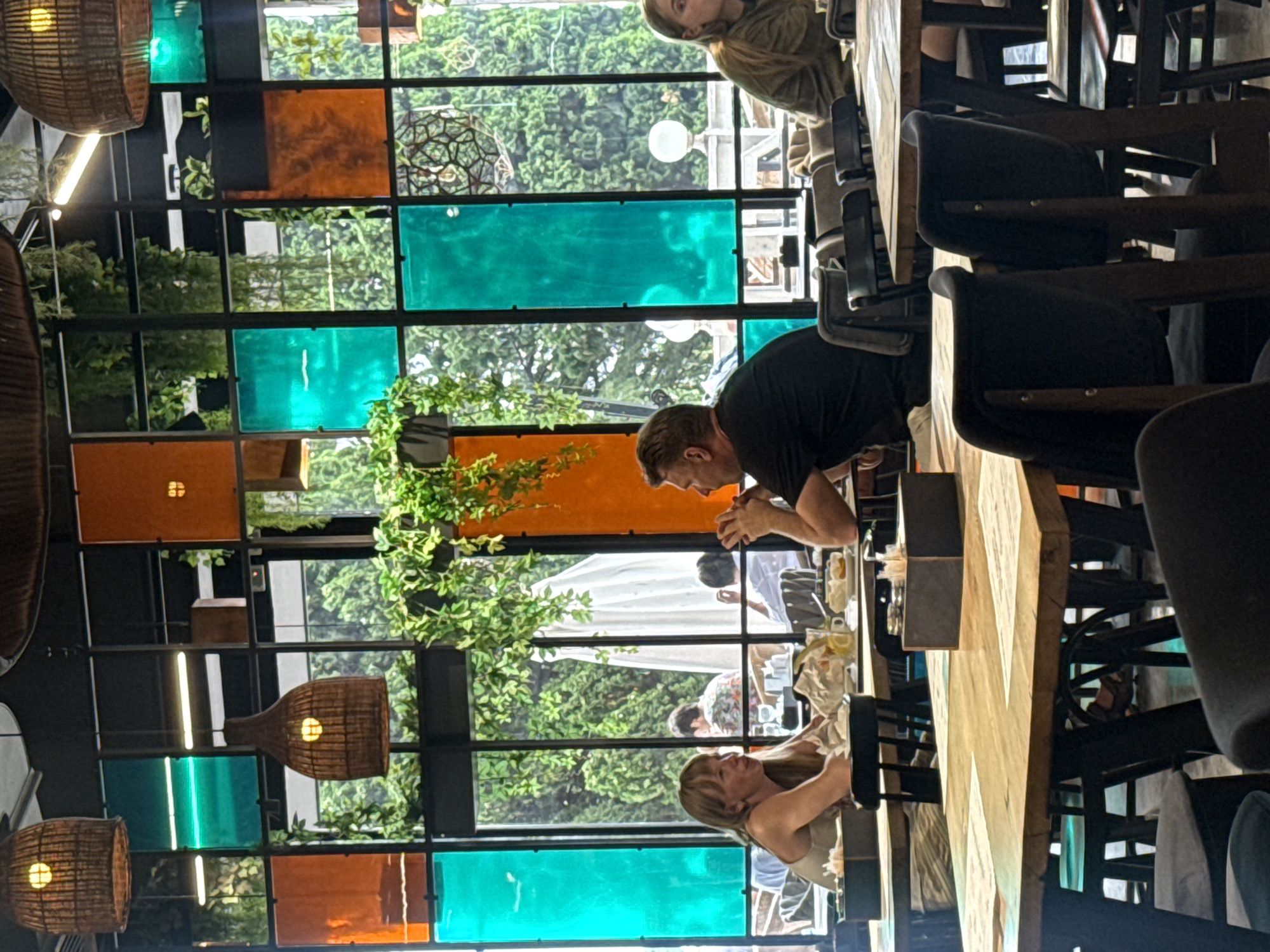 The image depicts the interior of a restaurant with a large window overlooking a lush, green outdoor area. The window is segmented with black frames and filled with alternating colored glass panels in shades of orange and teal, creating a vibrant, stained-glass effect. Several hanging wicker lamps with internal lights are visible from the ceiling.
In the foreground, a wooden table is set with papers, possibly menus, and a black box holder for napkins or cutlery. Several dark chairs surround the table.
A woman with blonde hair is seated at a table on the left, wearing a light-colored top and looking to her left. A man with short, light-brown hair and wearing a black t-shirt is seated opposite her, looking towards his left with his hands clasped. They appear to be engaged in conversation.
In the background, through the colored glass and the outdoor greenery, other patrons can be seen seated at tables. One person in the far background is wearing a brightly patterned shirt.
The overall ambiance suggests a modern, stylish restaurant with a focus on natural light and decorative elements. The location is identified as Gorod Yalta, Ukraine.