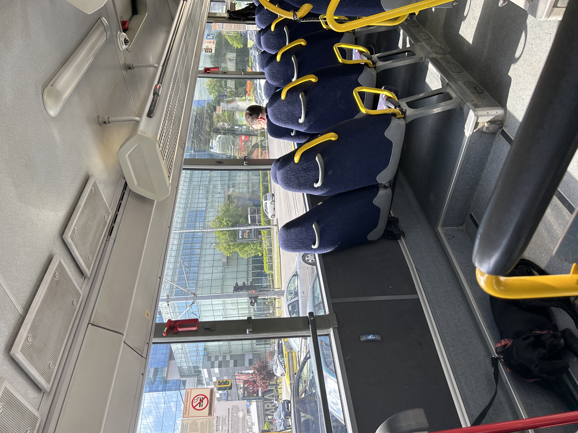 The image depicts the interior of a bus. A row of blue seats with yellow handholds lines the right side of the frame. A passenger is visible in one of the seats. Through the bus window, a modern building with glass facade is seen. Various cars are visible on the street. On the bus window are a "No Smoking" sign and advertising stickers. A black dog is lying on the floor near the front of the bus. The setting appears to be in an urban environment.