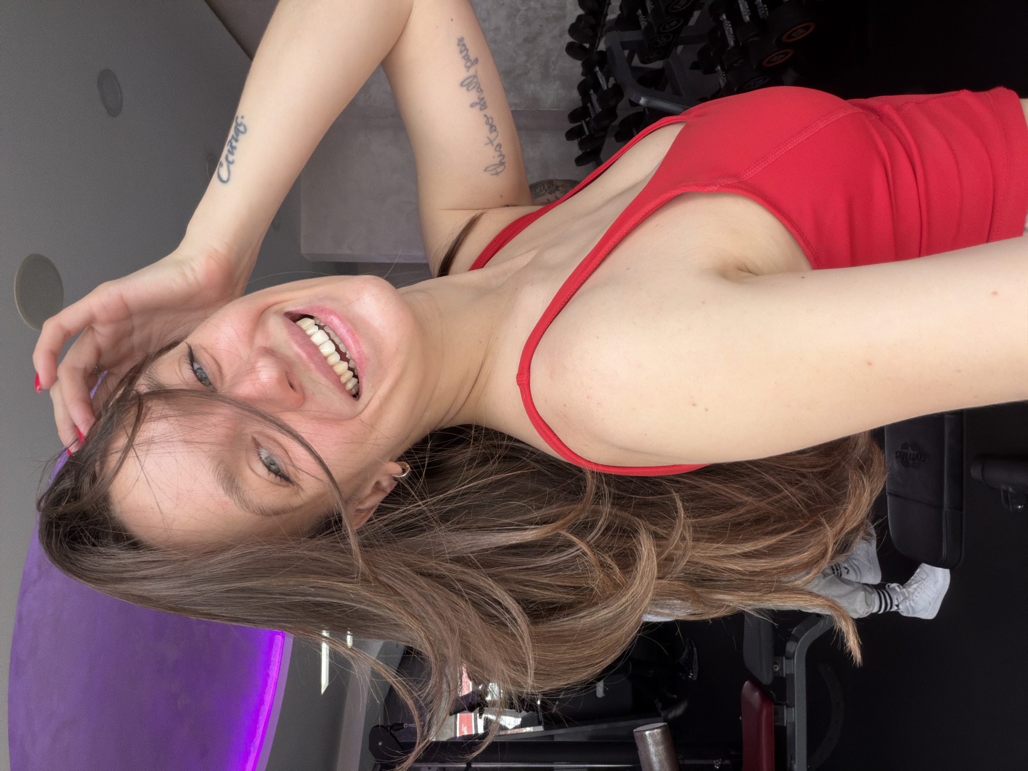 The image depicts an indoor fitness facility, specifically a gym, located within the city of Budva, Montenegro. The specific name of the facility is not discernible from the image.
The foreground is dominated by a female subject positioned off-center to the left, looking directly at the camera. She has long, brown hair styled with volume, with some strands falling across her face. Her eyes are light blue-green, and she is smiling broadly, revealing her teeth. Her right hand is raised, with fingers touching her hair near her forehead, exposing red-painted fingernails. She wears a red scoop-neck athletic tank top. Visible tattoos include Cyrillic script "Сейчас" (meaning "Now") on her right inner forearm and English script "this too shall pass" on her left inner bicep/upper arm. A gold-colored hoop earring is visible in her right ear.
The background elements confirm the gym setting. To the right of the primary subject, a multi-tiered rack of dumbbells, varying in size and weight, is visible. To the left, behind the subject's hair, a padded exercise bench with red and black upholstery is partially visible. Further back, obscured by the primary subject's hair, the lower torso and legs of a second individual are visible. This person is wearing light gray athletic pants with dark stripes down the side, white athletic shoes with dark striped socks, and appears to be seated or standing near equipment. The flooring is dark, likely black gym matting. The ceiling features recessed circular light fixtures, and a portion of a purple-lit decorative ceiling element is visible in the upper left. The walls are a light, neutral gray color.