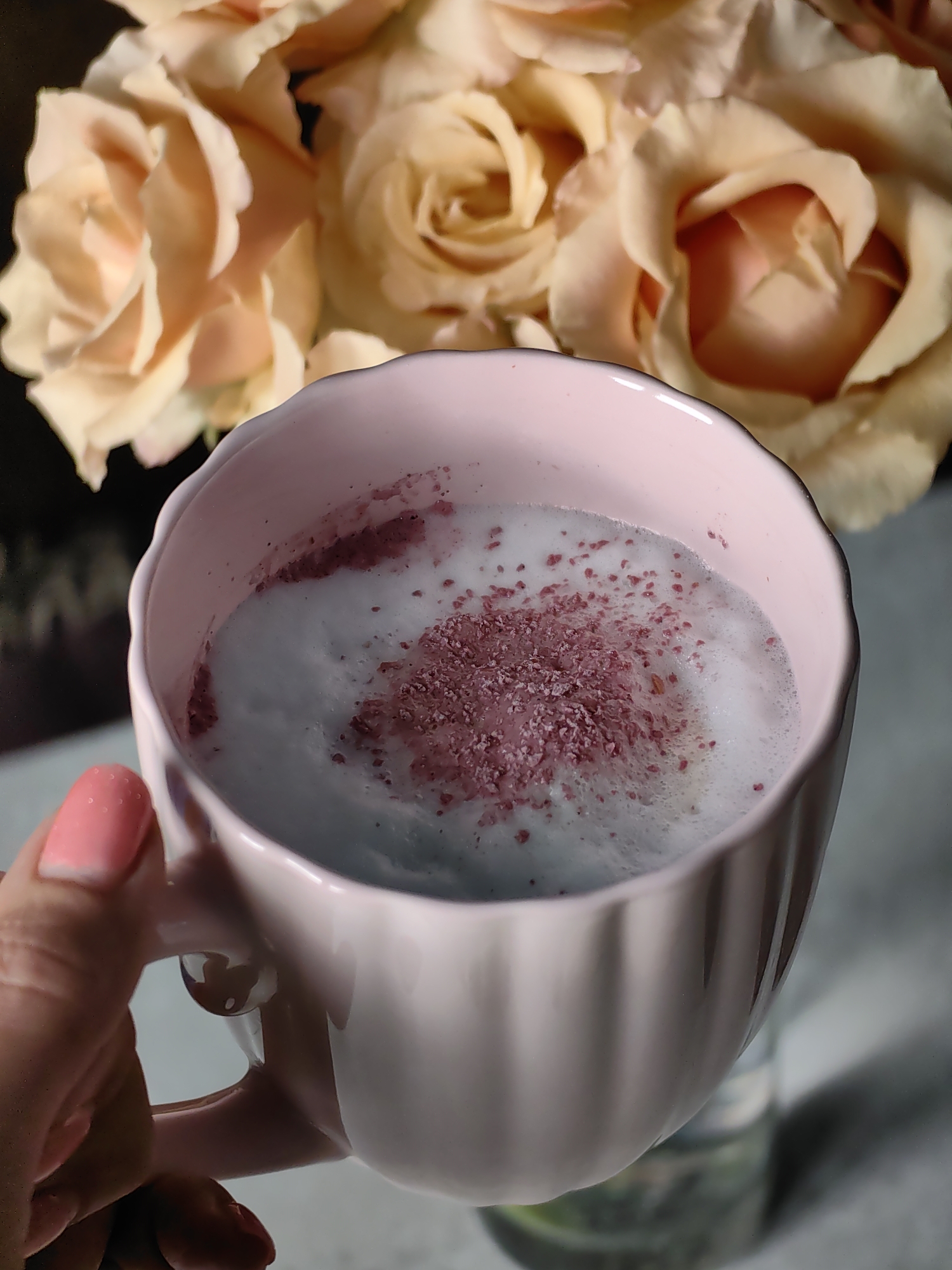 In Fanipal, Belarus, a close-up shot captures a light pink mug filled with a frothy, light blue beverage, possibly a latte or smoothie. The drink is sprinkled with fine, reddish-purple powder. A hand, with a light pink nail polish on its index finger, is holding the mug. In the blurred background, a bouquet of light peach-colored roses adds a soft, floral touch to the scene. The lighting is soft, highlighting the textures of the drink and the flowers.