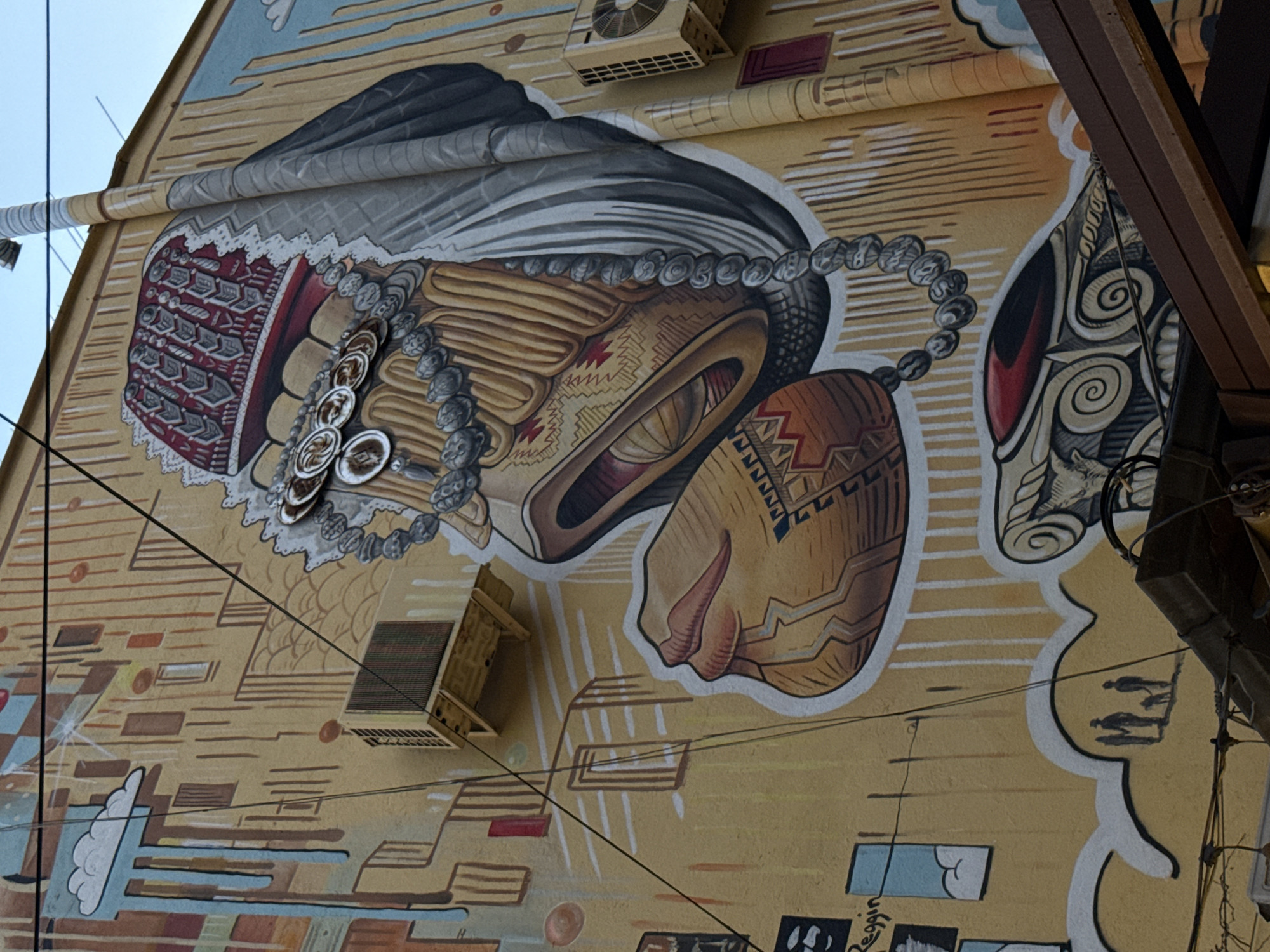 This image captures a large, intricate mural painted on the side of a building under a clear blue sky in Yerevan, Armenia. The central element of the mural is an abstract, stylized figure or entity presented against a tan background marked with vertical lines and geometric shapes suggesting urban structures.
At the top, a prominent red and grey headdress or crown with ornate, segmented patterns is depicted. Below this, a large, multi-strand necklace made of connected silver-colored medallions or coins drapes across the central facial area, descending into a long string of grey, spherical beads that extends downwards towards the lower right. A distinct, open, lip-like feature, rendered in browns and golds, is visible below the coin necklace. A flowing section of grey drapery extends from the upper right, partially covering the figure. Below the central figure, a separate, stylized brown and orange object with geometric patterns and a pronounced lip-like form is present. At the bottom of the mural, a grey and white architectural element featuring swirling volute designs is visible.
The background of the mural includes various painted elements: in the upper left, a white cloud with blue, dripping streaks; in the lower left, small framed images, one bearing the text "Reggin" and another showing a cloud; and below these, black silhouettes of three people—two adults and one child—accompanied by a dog, walking and casting shadows.
Interspersed with the mural are several real-world urban objects. Two yellowish-beige outdoor air conditioner units are mounted on the wall: one on the upper left and another on the upper right, the latter displaying a fan grille. A large, corrugated pipe, painted to match the mural's aesthetic, runs vertically along the right side of the building. Multiple black electrical wires crisscross the frame diagonally. A dark brown structural beam or awning partially covers the lower portion of the mural at the bottom of the image.