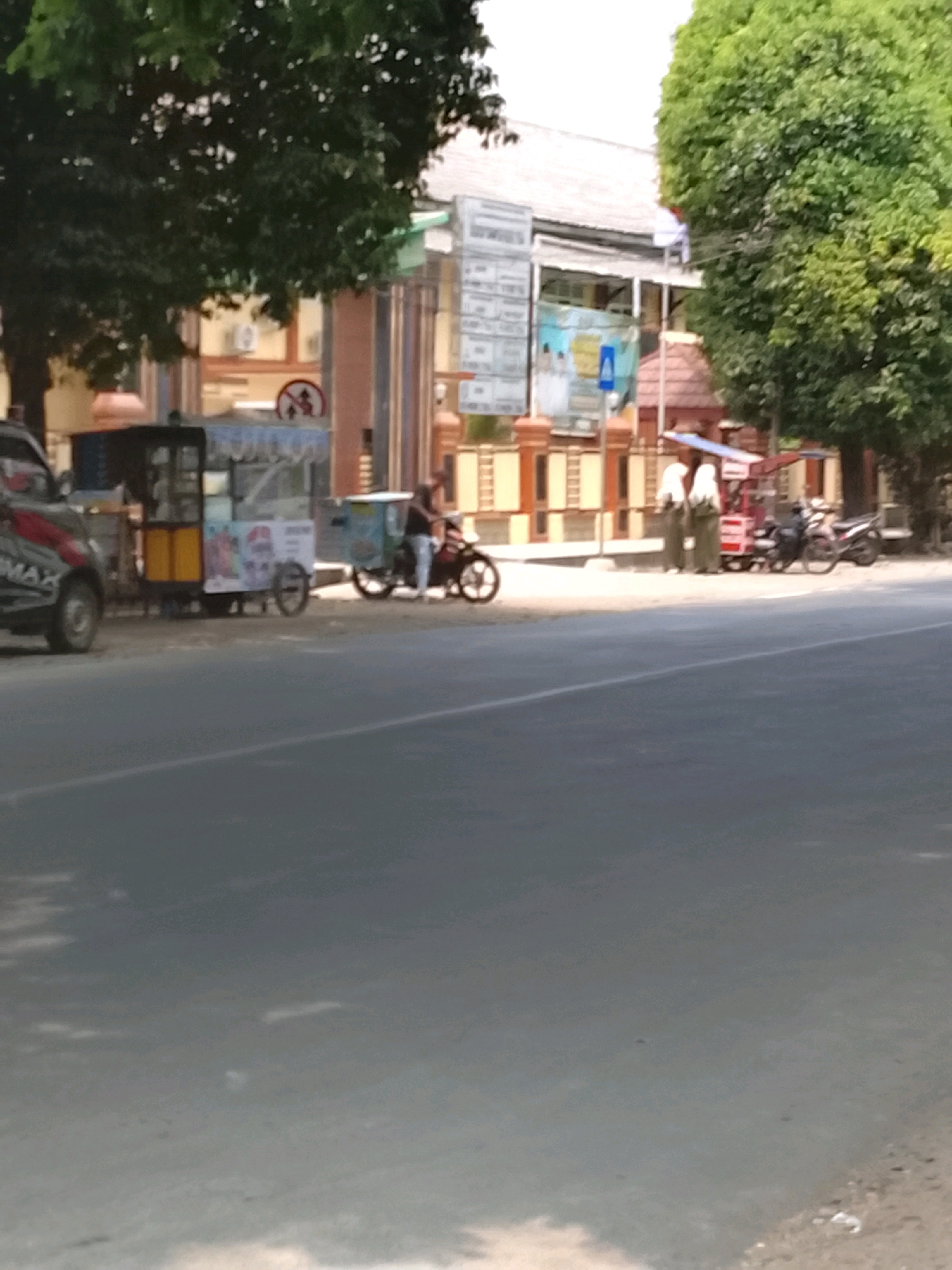 In Slawi, Indonesia, a street scene unfolds under a bright sky. On the left, a parked dark-colored vehicle with red accents is visible, its front tire turned. Adjacent to it is a colorful food cart with yellow paneling and a white top. Further down the street, a person on a motorcycle is stopped near another, smaller food cart. To the right, two individuals in light-colored attire stand near a red stall with a blue awning. Behind them, a white and gray building with a tiled roof is partially obscured by lush green trees. The street itself is paved, with a visible white line marking the lanes. The overall impression is one of a bustling roadside in an Indonesian city.
