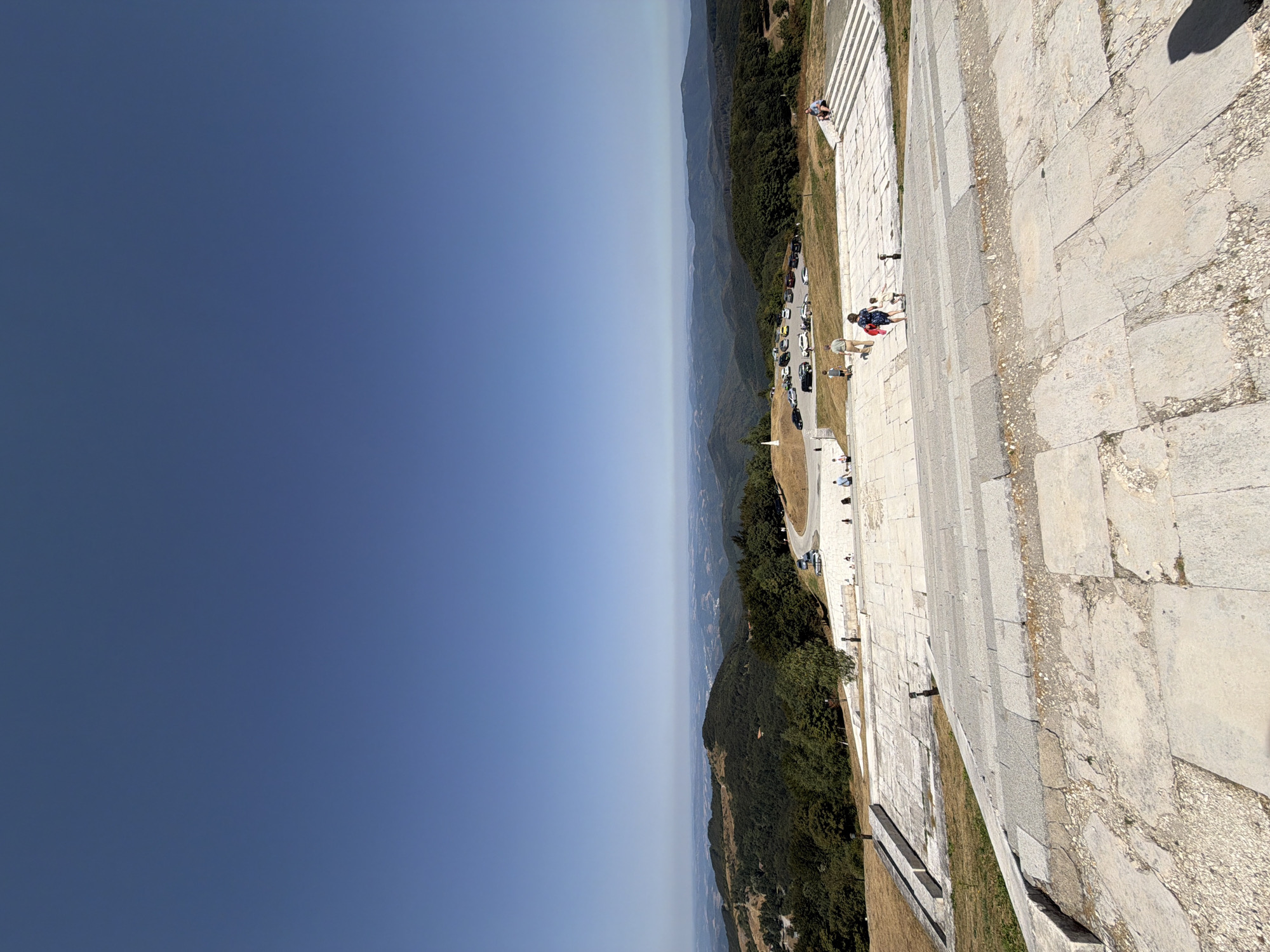 The image captures a wide, panoramic view from a stone staircase overlooking a vast mountainous landscape under a clear blue sky. The foreground is dominated by the textured, weathered stone steps of the staircase, showing signs of age and wear.
In the middle ground, a paved area features several parked cars and a small group of people. Some individuals are walking, while others are seated or standing. A white monument or marker is visible in the distance on the left. Beyond this, rolling hills covered in dense green forest stretch towards the horizon.
The background is a hazy, layered vista of distant mountains and valleys, fading into the atmospheric perspective. The scene suggests a popular viewpoint or memorial site in a mountainous region. The location is identified as the Buzludzha Monument, a significant historical landmark situated in the Shipka Pass, Bulgaria.