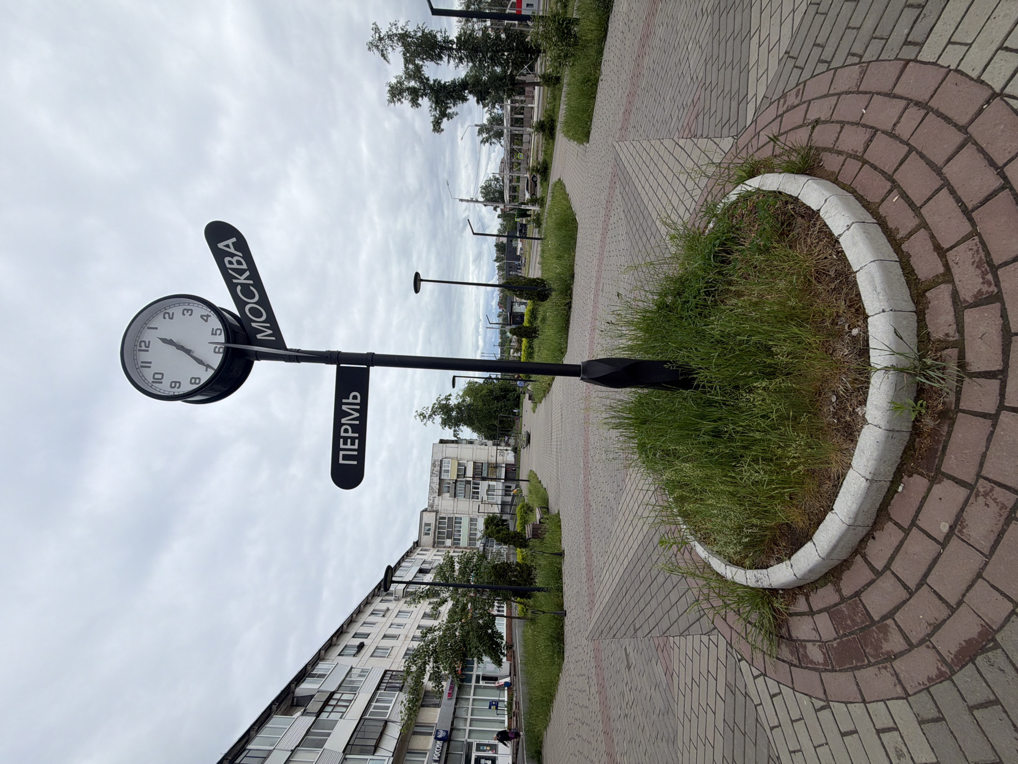 The image shows a street clock in the city of Chaikovsky, Russia. The clock has two signs pointing to "Moscow" and "Perm." It is placed on a black metal pole surrounded by a circular flowerbed filled with grass. The flowerbed is edged with white concrete blocks. The ground is paved with red and gray bricks. In the background, there are residential buildings and trees. The sky is overcast.