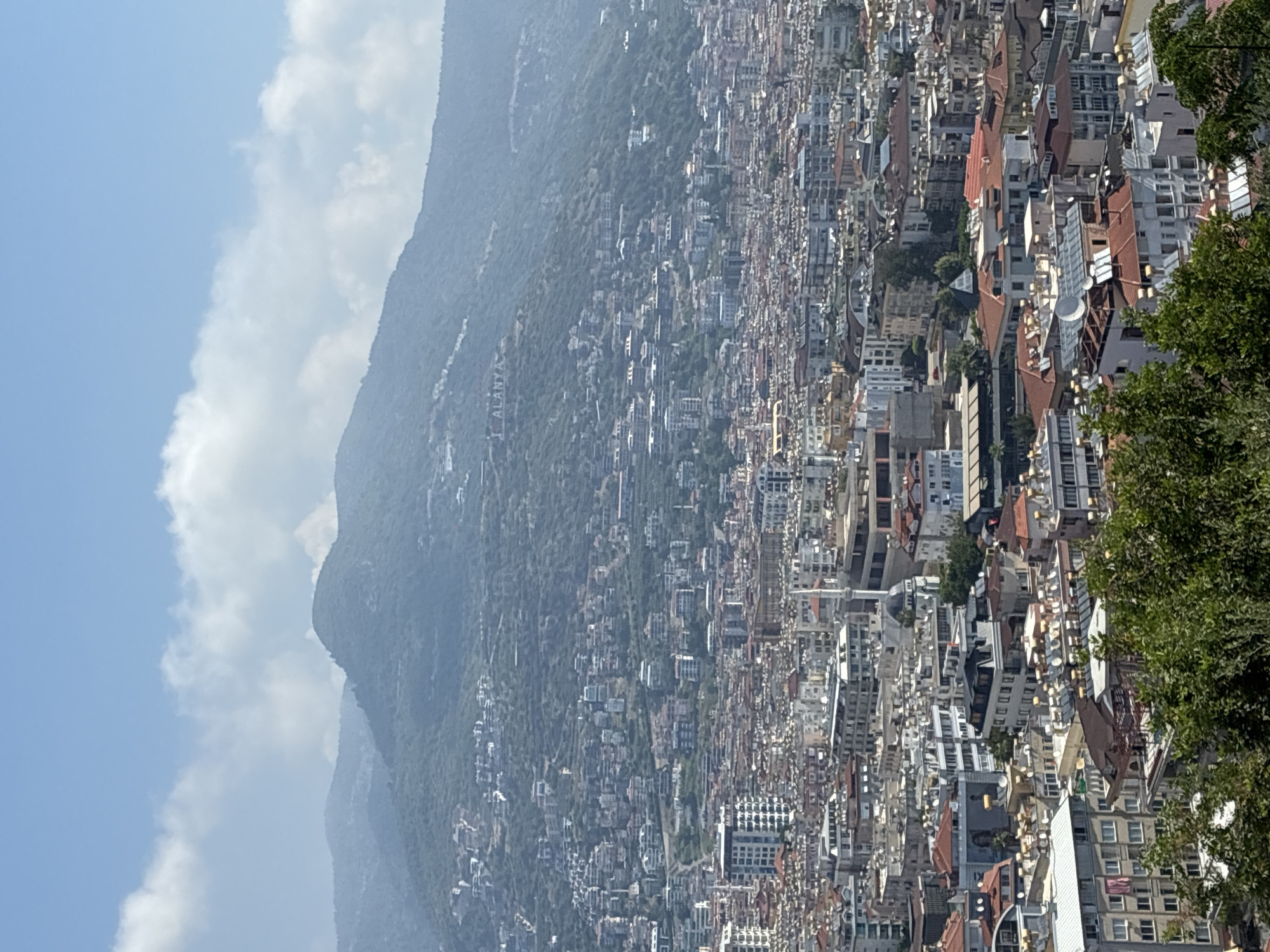 This is a high-angle, medium-shot, eye-level photograph of the city of Alanya, Turkey, on a bright, sunny day with scattered clouds. The foreground is dominated by lush green trees. In the middle ground and background, a dense urban landscape unfolds, characterized by a multitude of buildings with varying architectural styles, colors, and roof types, including many with terracotta-colored roofs. Interspersed among the buildings are solar water heaters, satellite dishes, and the distinct white minaret of a mosque.
Further in the background, a large, tree-covered mountain rises majestically. The slopes of the mountain are dotted with more buildings, creating a sense of the city extending upwards. Near the top of the mountain, a large, white inscription is visible on the hillside, appearing to spell out "I ALANYA". The sky above is a clear blue, with wispy white clouds. The overall impression is that of a bustling coastal city nestled against a dramatic natural backdrop. There are no people clearly visible in the frame.