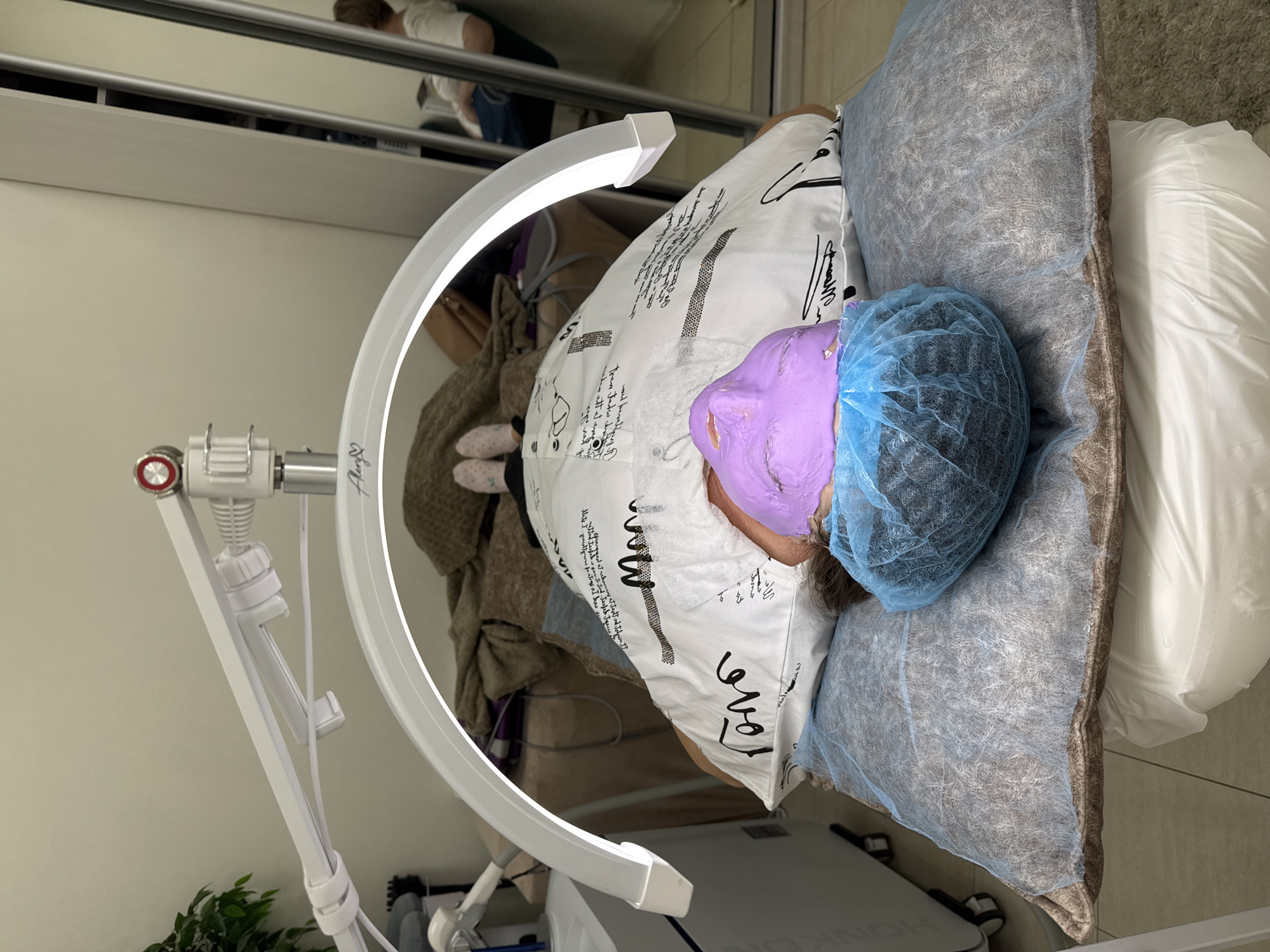 A person wearing a blue surgical cap and a purple facial mask lies on a bed in what appears to be a spa or salon setting. The person rests on a pillow covered in a white fabric with black writing and designs. The bed is covered with a light blue disposable sheet. A white ring light stands to the left of the bed, illuminating the scene. In the background, a mirrored closet is visible. The environment seems clean and professional.