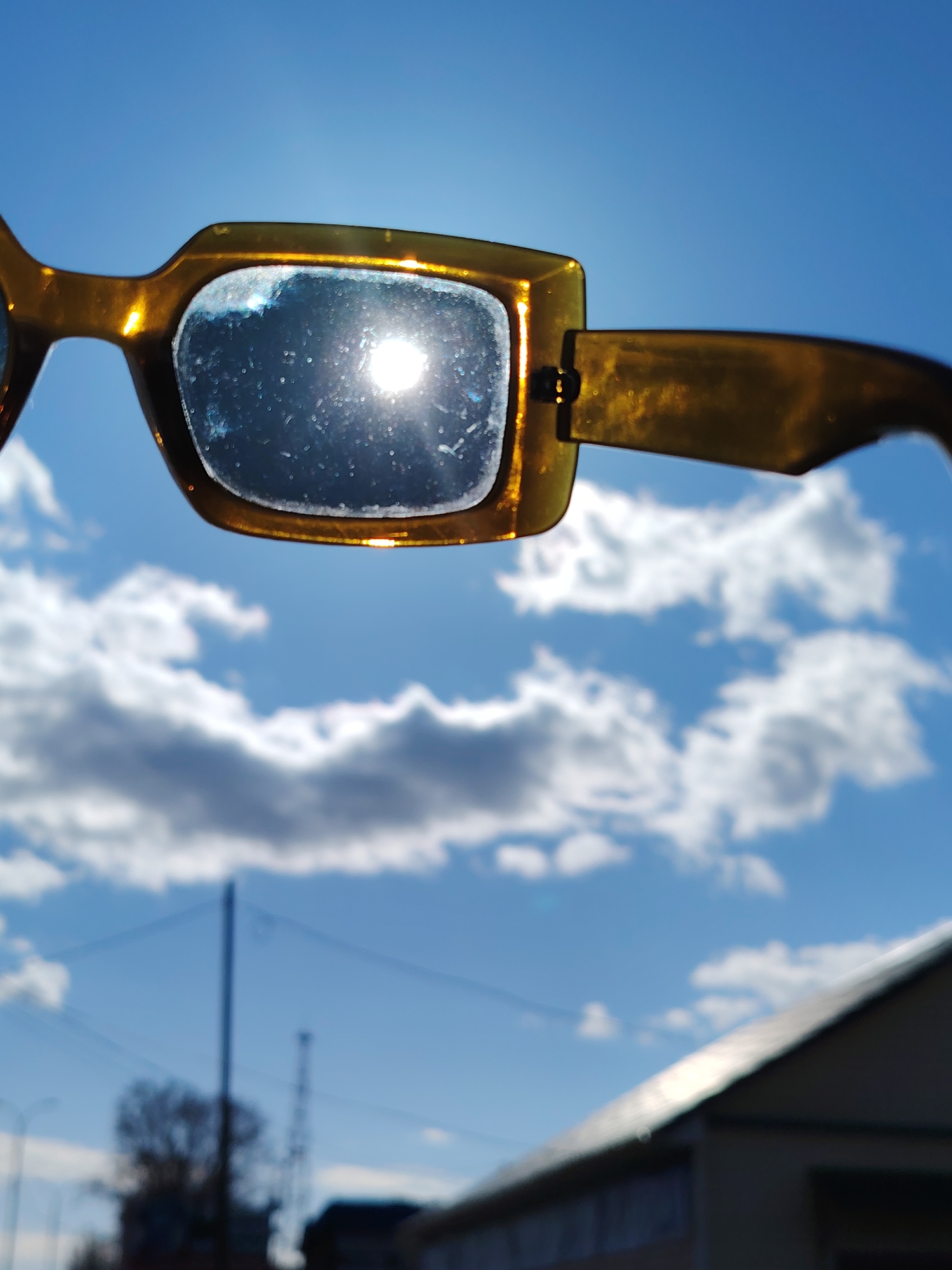 The image shows a pair of eyeglasses held up to the sky on a sunny day. The sky is bright blue with puffy white clouds. The sun is reflected in one lens of the eyeglasses. In the background, there is a building with a sloping roof, a tree, and what appears to be a utility pole. The location appears to be in an urban environment. The city of Chaikovsky, Russia is not identifiable with the information provided in the image.
