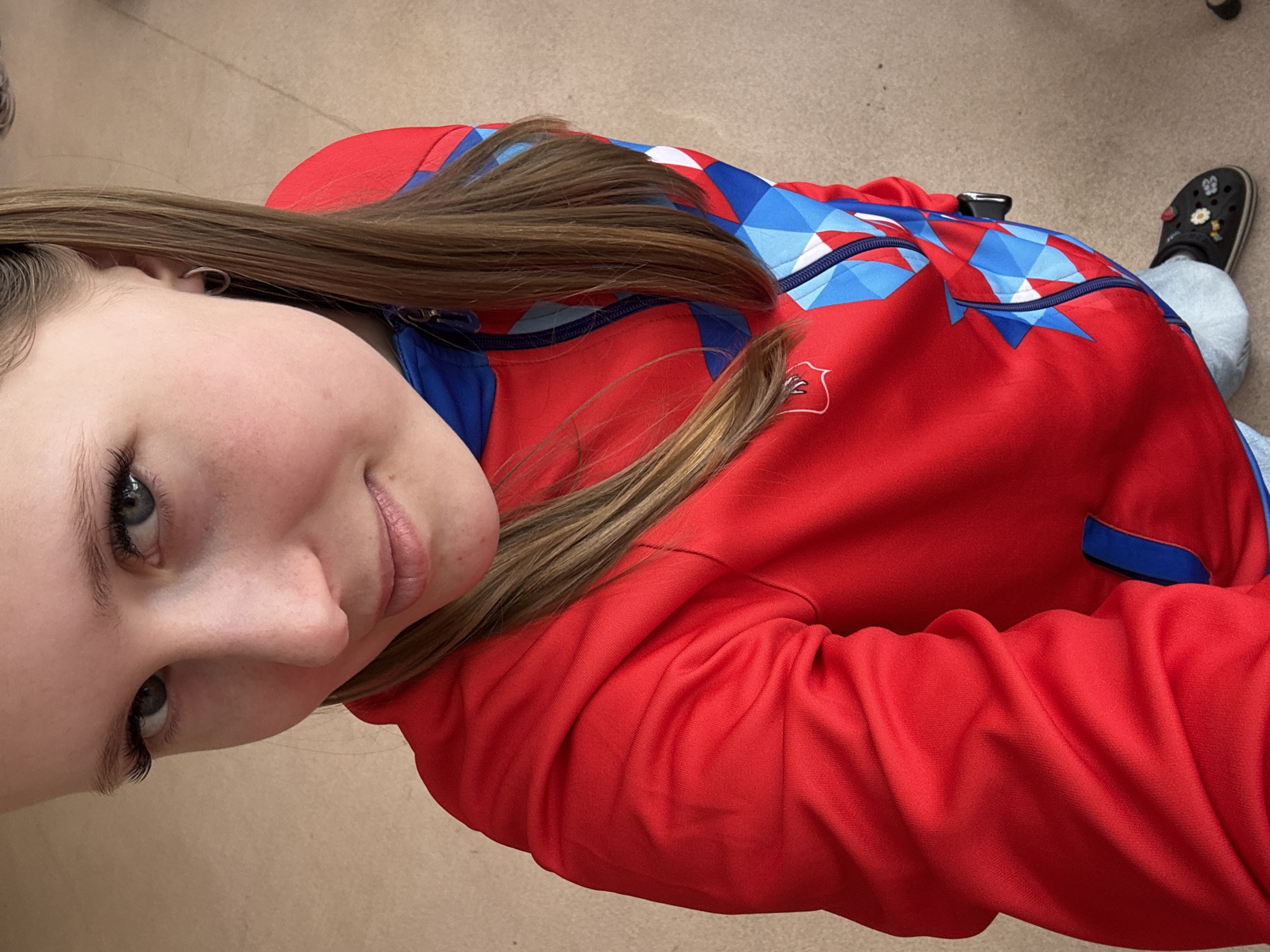 A close-up, rotated image shows a young woman with light brown hair and grey-blue eyes. She is wearing a red jacket with blue and white geometric patterns on the upper chest and shoulders. A small emblem is visible on the lower left chest. The jacket has blue trim. The woman is wearing what appears to be a simple hoop earring. The background is a neutral beige color, likely a floor. In the background, a person is wearing grey jeans and black Crocs with flower embellishments. Based on the jacket design, this photo was likely taken in the city of Usolye, Russia.