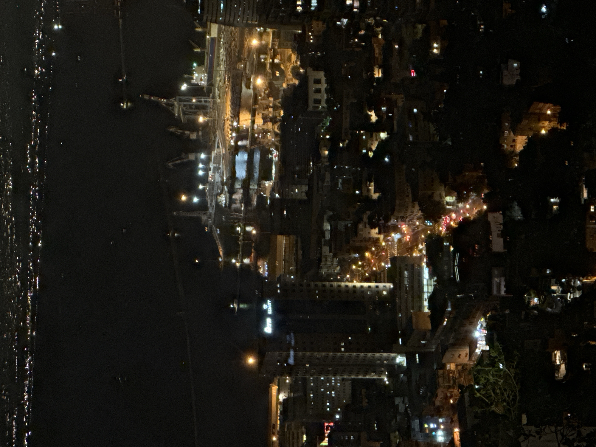 The image is a nighttime aerial view of the city of Haifa, Israel, looking towards the port and the Mediterranean Sea. The foreground is dominated by dark, silhouetted trees and buildings. In the mid-ground, there are illuminated buildings, including a tall, modern skyscraper with illuminated signage that reads "SEA CITY". The port area is visible with numerous cranes, ships, and bright lights reflecting on the water. The background shows the distant lights of the city and the expanse of the dark sea. The scene depicts a bustling urban and industrial environment at night.