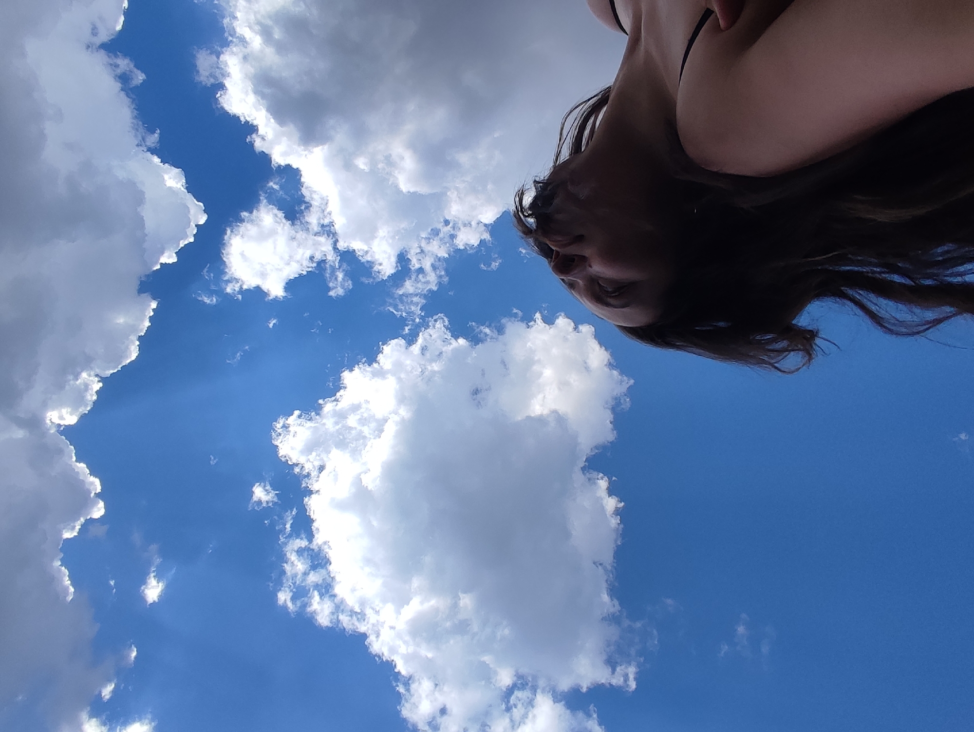 The image shows a low-angle view of a person against a blue sky with scattered white clouds. The person's head and upper torso are visible, with long dark hair. The background is composed primarily of blue sky and white, fluffy cumulus clouds. The location is not evident from the image content. Фоки, Russia is not evident from the image content.