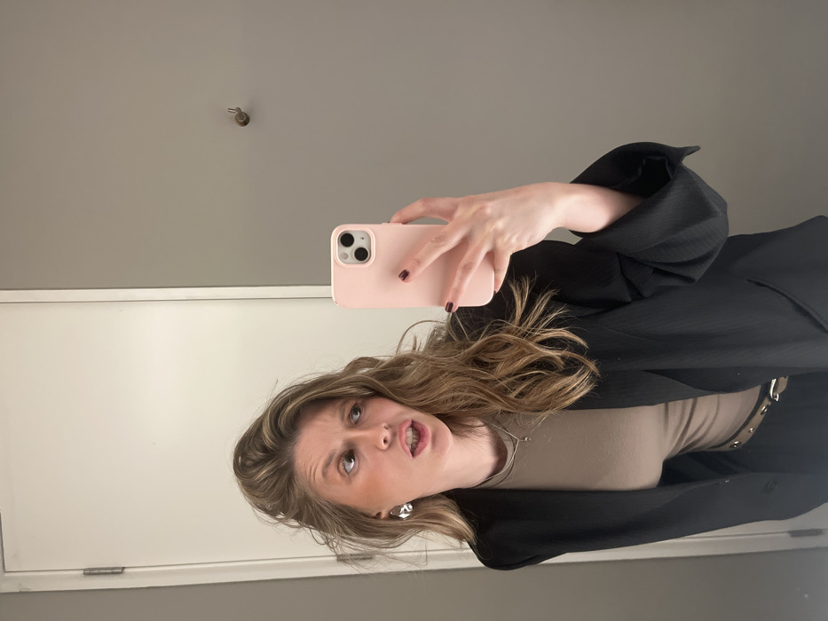 A person with blonde, wavy hair holds a pink iPhone in their right hand, presumably taking a selfie. The person wears a beige top under a black blazer with rolled-up sleeves. The background features a white door and a gray wall with a small hook. The person's facial expression includes slightly parted lips and raised eyebrows. They also wear silver hoop earrings and dark nail polish.