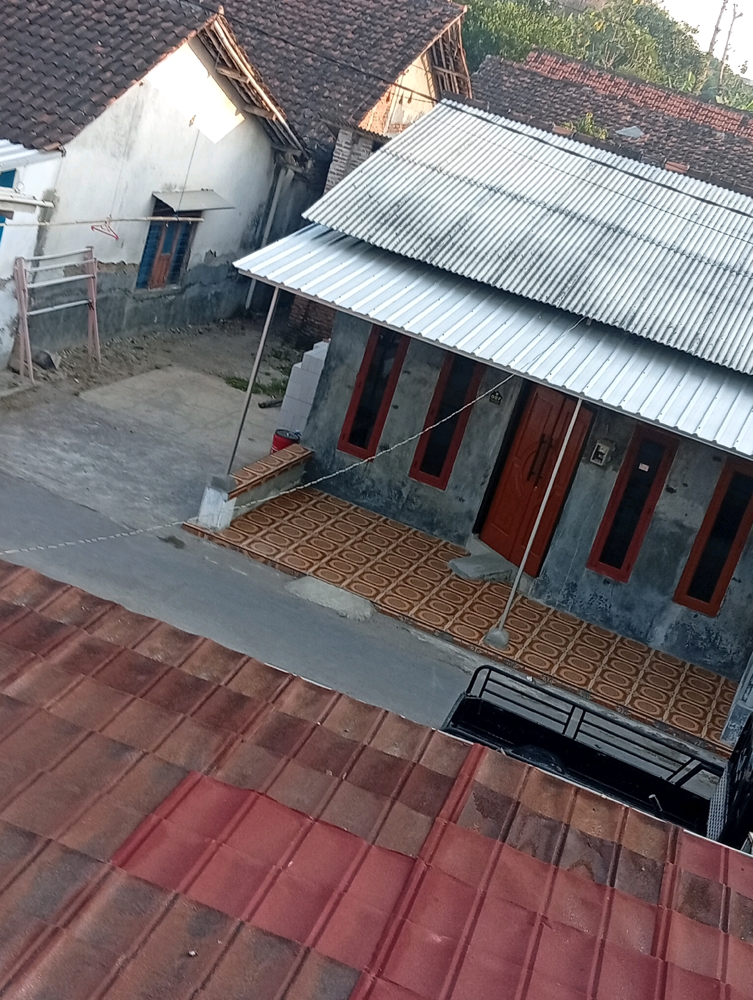 The image captures a street scene in Kebandingan, Indonesia, featuring a row of houses and their roofs. In the foreground, a weathered, reddish-brown tiled roof dominates the lower portion of the frame. Further back, a street is visible, separating the foreground roof from a small, gray-walled house with a corrugated metal roof. This house has a prominent red door and multiple windows with red frames. In front of the house, a patterned tiled patio is laid out. To the left of this house, another, older house with a white-painted wall and a dark tiled roof is partially visible, showing a window with a wooden shutter and a metal drying rack outside. A small black truck is parked to the right of the house with the corrugated metal roof, its railing visible. The environment appears to be a residential area with a somewhat aged infrastructure, typical of many neighborhoods in Indonesian cities.