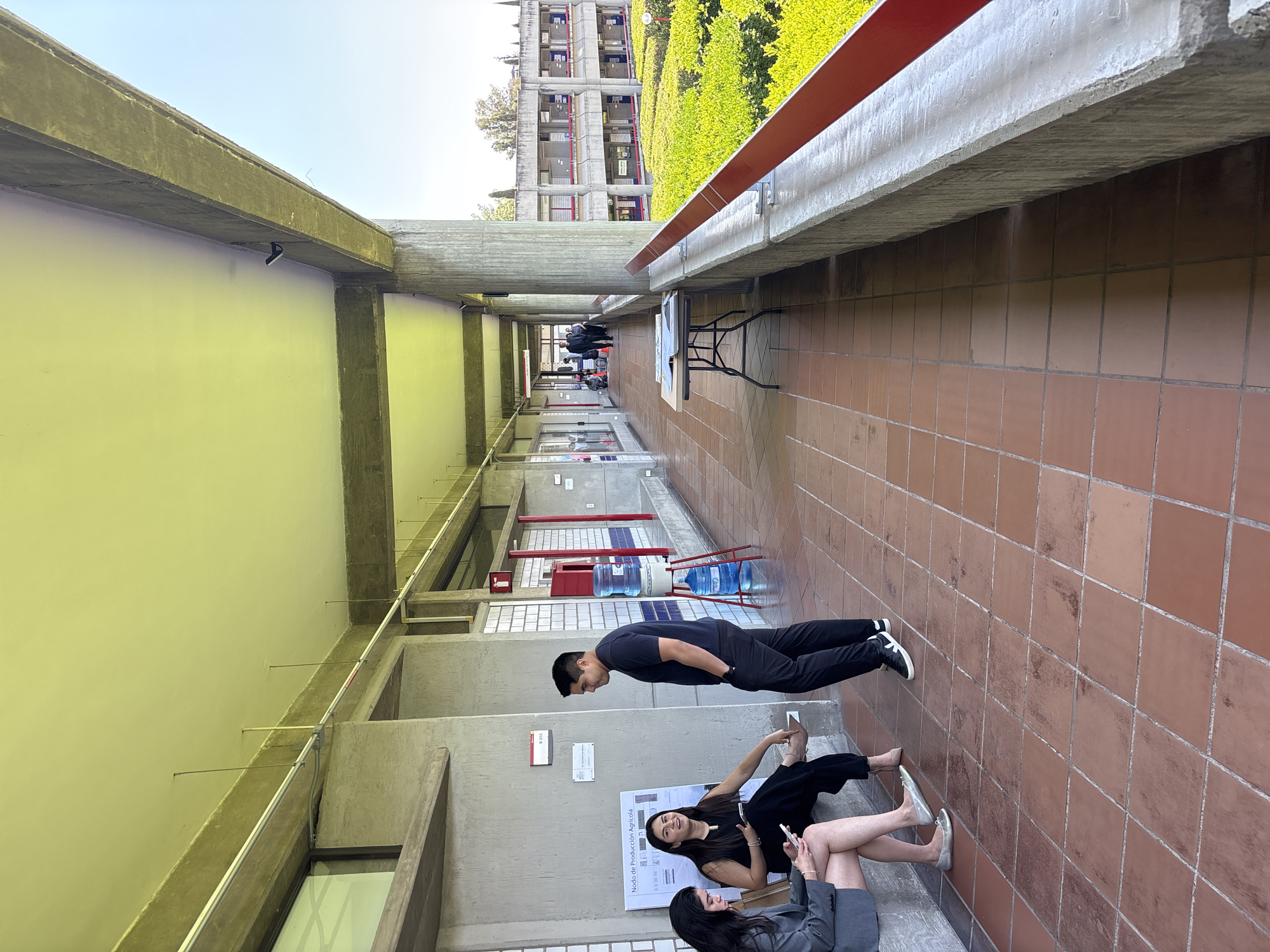 A long outdoor hallway with red tile floors is lined with concrete walls and ceilings. The left wall is painted yellow, and the right wall has a red rail running along its top. A man in black clothes is standing towards the bottom of the frame, with two women seated near him, one in black and one in a gray suit. They are near a poster on the wall. In the background, a building covered in green foliage is visible. There is also an empty table with a black frame. It appears that the image was taken at the Benemérita Universidad Autónoma de Puebla (BUAP) in Puebla, Mexico, as the architecture and foliage on the buildings are consistent with the architectural design.