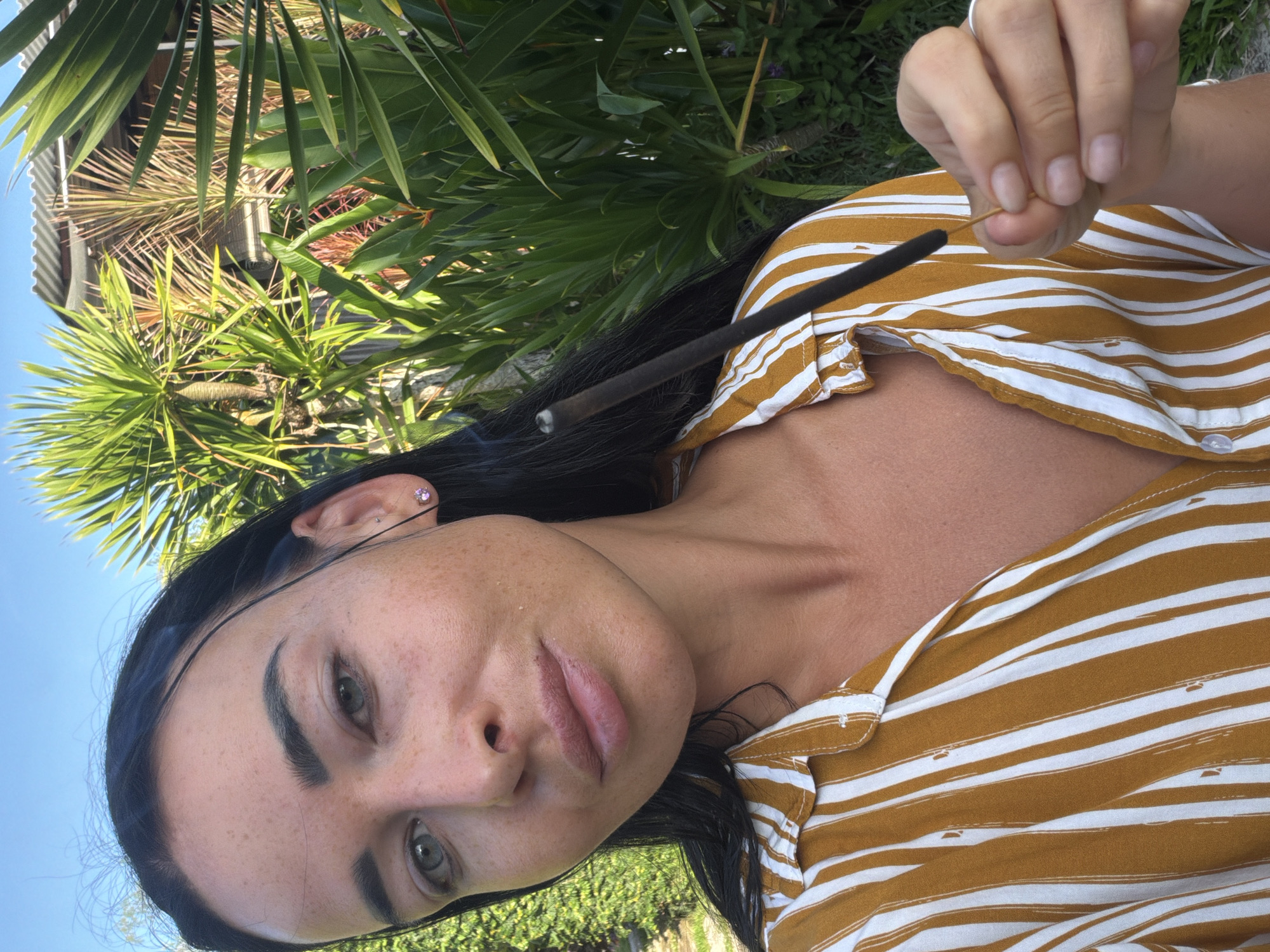 A woman holds a lit incense stick in her right hand in an outdoor setting. The woman has dark hair, fair skin with freckles, and is wearing a mustard-colored and white striped shirt. Her left ear has a purple earring. The background includes greenery, specifically palm trees and a grassy area. The photo appears to be taken during the day with natural light. The location is Kutuh, Indonesia.