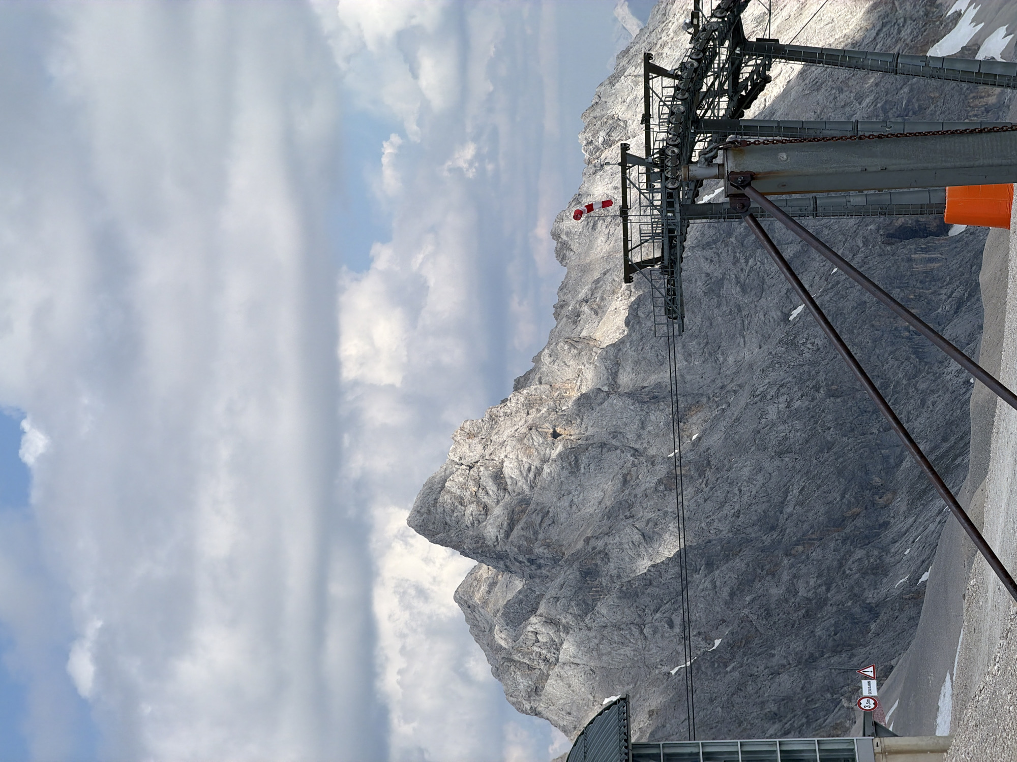 This media file captures a dramatic mountain landscape under a partly cloudy sky, likely during the daytime given the lighting. The primary subjects are the imposing, rugged grey mountains that dominate the scene, with patches of snow visible on their slopes. In the foreground, a section of a cable car infrastructure is prominent, featuring a large metal pylon with a complex system of pulleys and cables. An orange object, possibly a protective covering or part of the structure, is attached to the pylon. A red and white striped windsock is also visible, attached to the cable car structure.
The overall setting is a mountainous area, suggested by the terrain and the cable car, which is commonly used for accessing such high altitudes. The activity is implied to be related to mountain transportation, likely a ski resort or a scenic viewpoint. The weather appears to be mixed, with bright sunlight casting shadows on the mountains and illuminated clouds drifting across the sky.
Several signs are visible in the lower right portion of the image, though they are small and partially obscured. One sign appears to have a warning symbol and text, and another circular sign indicates a speed limit of "3.5m". One sign has German text that reads "SEILBAHN", which translates to "cable car" or "funicular". The presence of these signs and the cable car suggests the location is a tourist or recreational area within the German Alps, consistent with the Garmisch-Partenkirchen context. The overall mood is one of natural grandeur and engineered human infrastructure coexisting in a challenging environment.