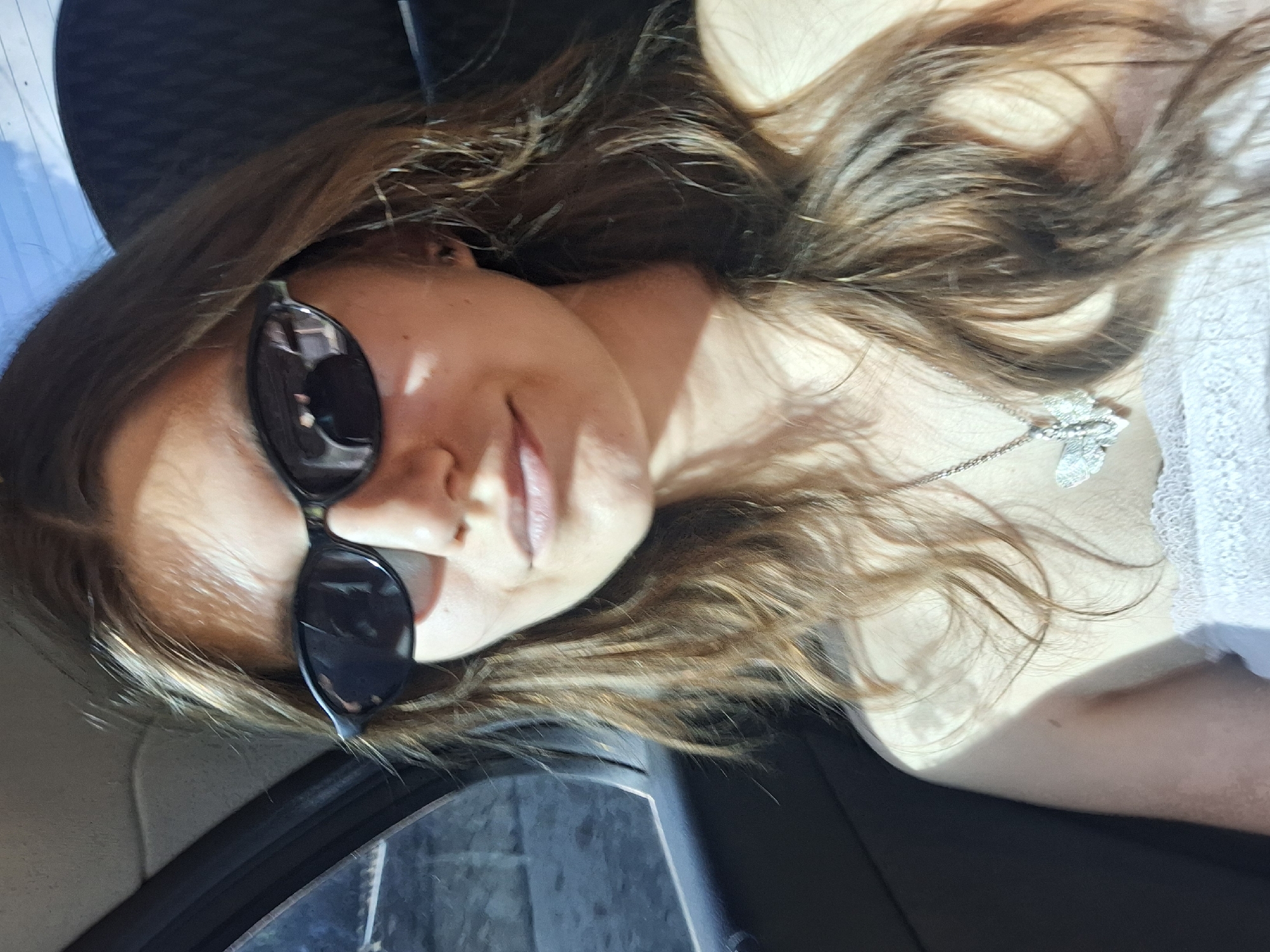 The media file is a close-up, horizontal shot of a young woman wearing sunglasses and a necklace. She is lying down, possibly in a car given the visible dark interior and window reflections. Her long, brown hair is spread out, with some strands catching the sunlight. The woman appears to be looking at the camera, with a slight smile on her face, suggesting a relaxed and perhaps candid moment. The lighting indicates it is daytime, with strong sunlight creating shadows on her face and hair. The overall mood is casual and possibly adventurous, given the sunglasses and the context of being on the move. There is no visible text in the image.