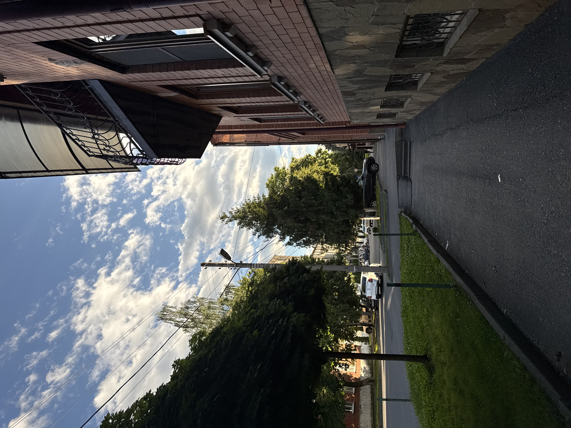 The image shows a sidewalk next to a brick building with a balcony. There are trees and cars on the street in the background. The sky is blue with white clouds. The scene appears to be in a residential area.