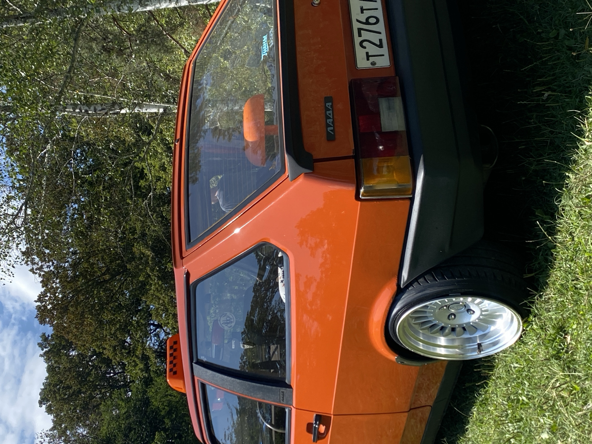 An orange Lada hatchback with a taxi sign on its roof is parked on a grassy area with trees and a blue sky with clouds in the background. The car has a custom silver rim on the rear passenger-side wheel. A license plate that reads "T276TA" is visible on the rear of the car. The scene appears to be outdoors, possibly in a rural setting or park, and not in the city of Bezymyannoe, Russia.