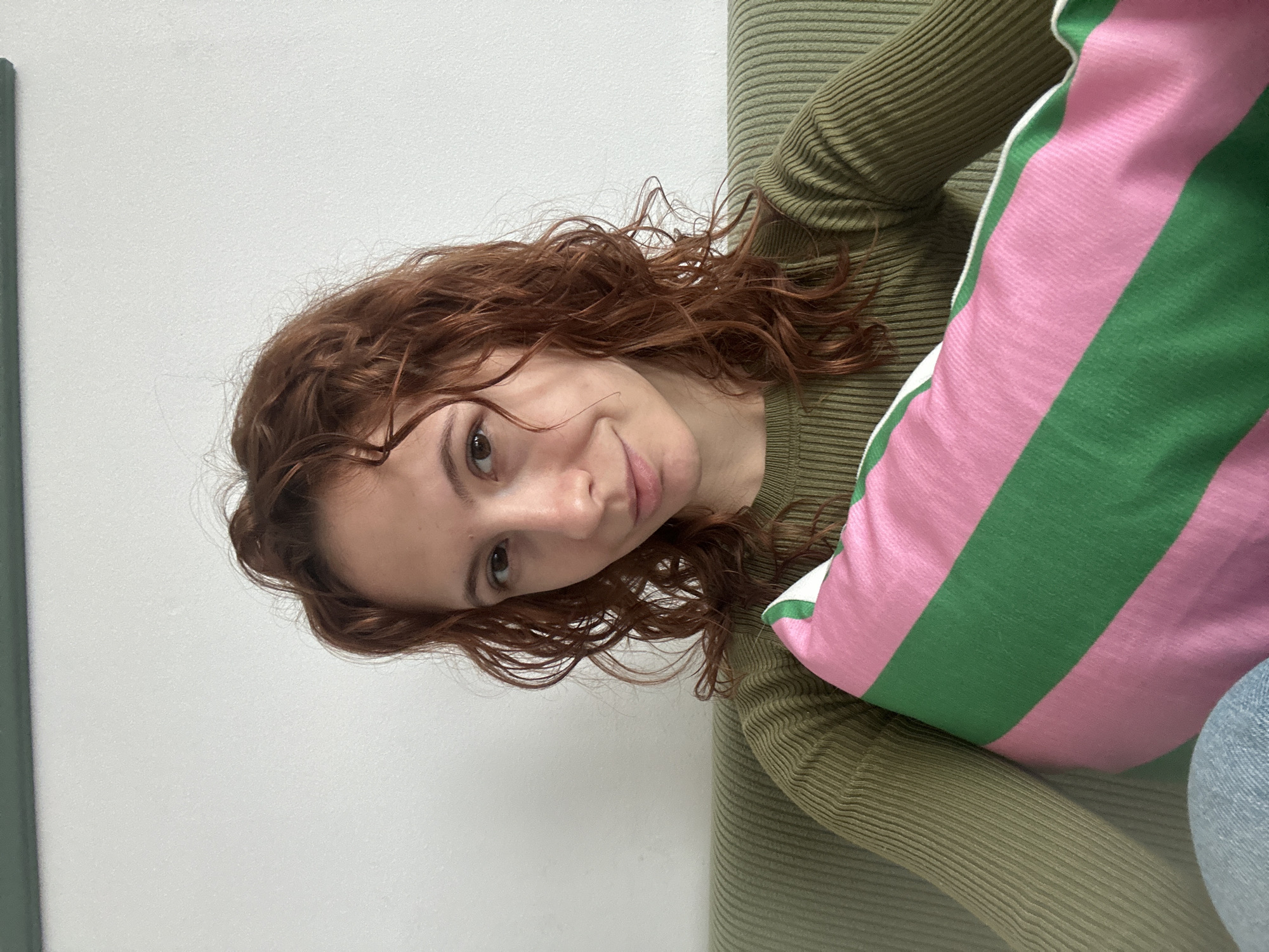A young woman with auburn curly hair is shown from the chest up, resting against a light green ribbed couch with a pink and green striped pillow. She is wearing a matching light green ribbed long-sleeved shirt. The background is a plain white wall. The location cannot be determined as Novi Sad, Serbia from the visual information.