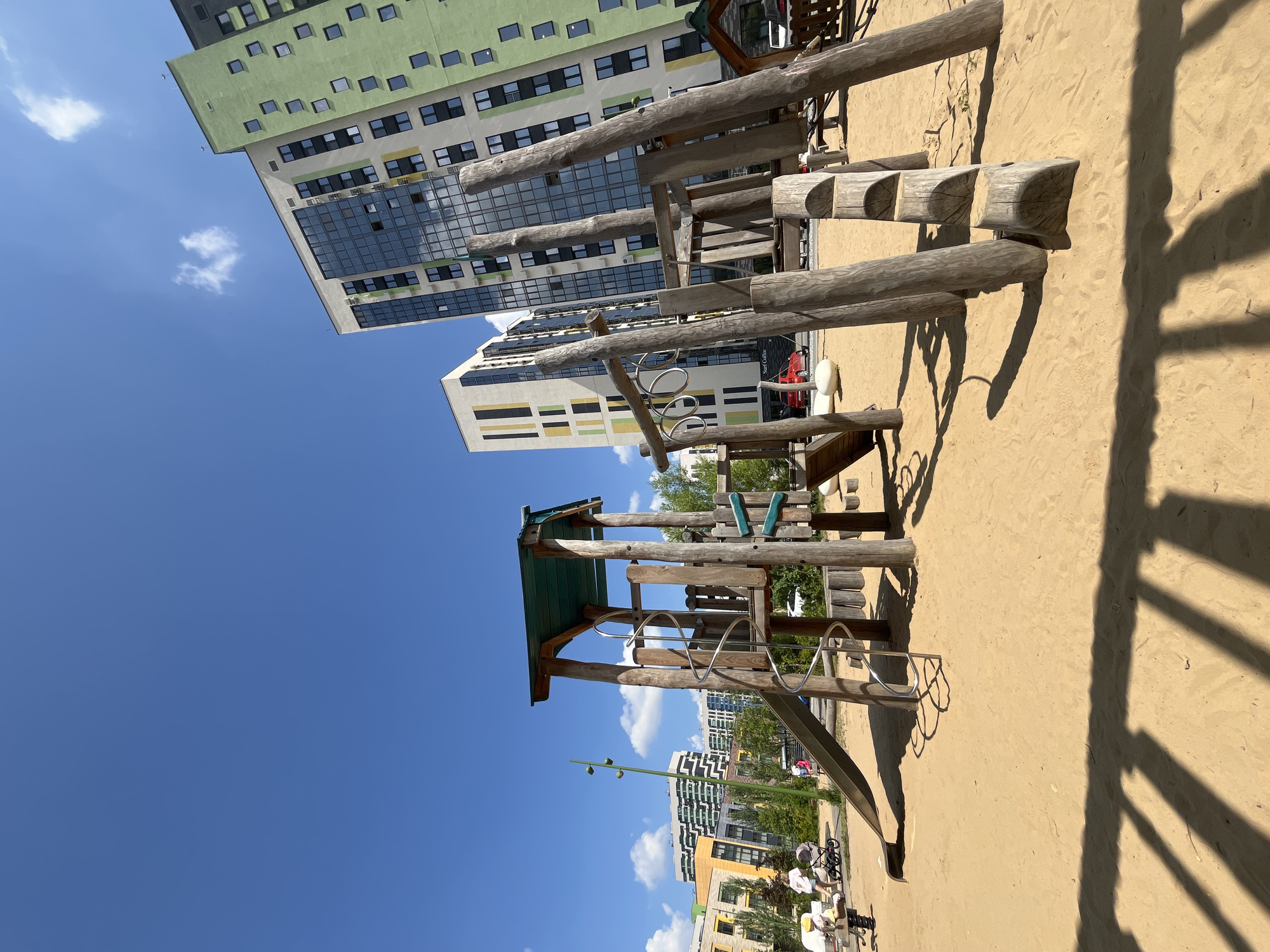 An outdoor wide-angle shot captures a modern urban playground with a wooden play structure in the foreground. The play structure, made of natural logs, features a slide, a spiral staircase, and a climbing area with horizontal logs acting as steps. The ground of the playground is covered in sand. In the background, several contemporary apartment buildings with colorful facades and large windows rise against a bright blue sky with scattered white clouds. A few trees are visible between the buildings and behind the playground. On the left side of the image, two adults and a child with a stroller are present, suggesting a family-friendly urban environment. The overall scene depicts a sunny day in a developed city, where residential architecture meets recreational spaces for children.