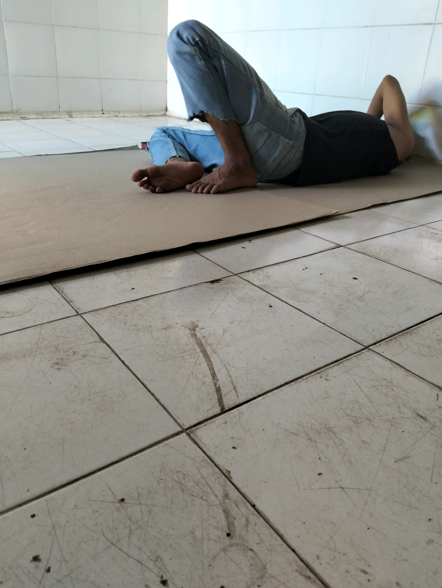 Two individuals are lying on the floor of a room with tiled flooring. The floor is dirty and appears to have scuff marks. The walls are white tiled. One individual is lying on their side, wearing a black t-shirt and blue jeans. Their legs are bent, and their right arm is raised and bent, resting near their head. Their face is not visible. The second individual is partially visible in the foreground, also lying on their side with their legs bent. They are wearing blue jeans, and their feet are visible, appearing bare. Both individuals are positioned on a piece of cardboard that is laid out on the floor. The scene is shot from a low angle, focusing on the lower bodies and the floor.