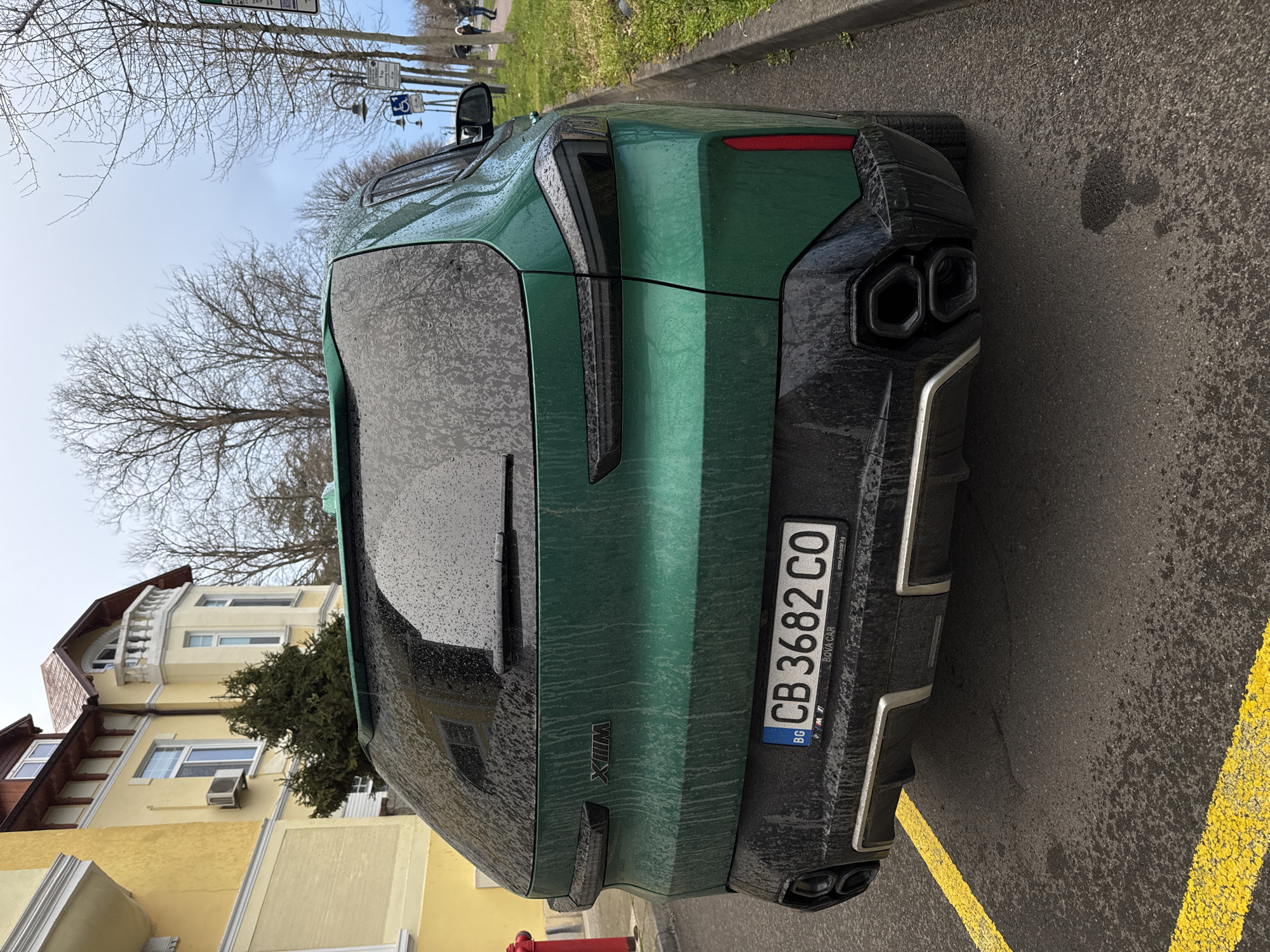 The image shows the rear of a green BMW XM parked on a street, with a Bulgarian license plate reading "CB 3682 CO". The car is wet, likely from rain. The background includes a multi-story yellow building and bare trees. The location is Burgas, Bulgaria based on the license plate region code "CB". There are yellow lines painted on the street. A fire hydrant is visible on the left side of the image.