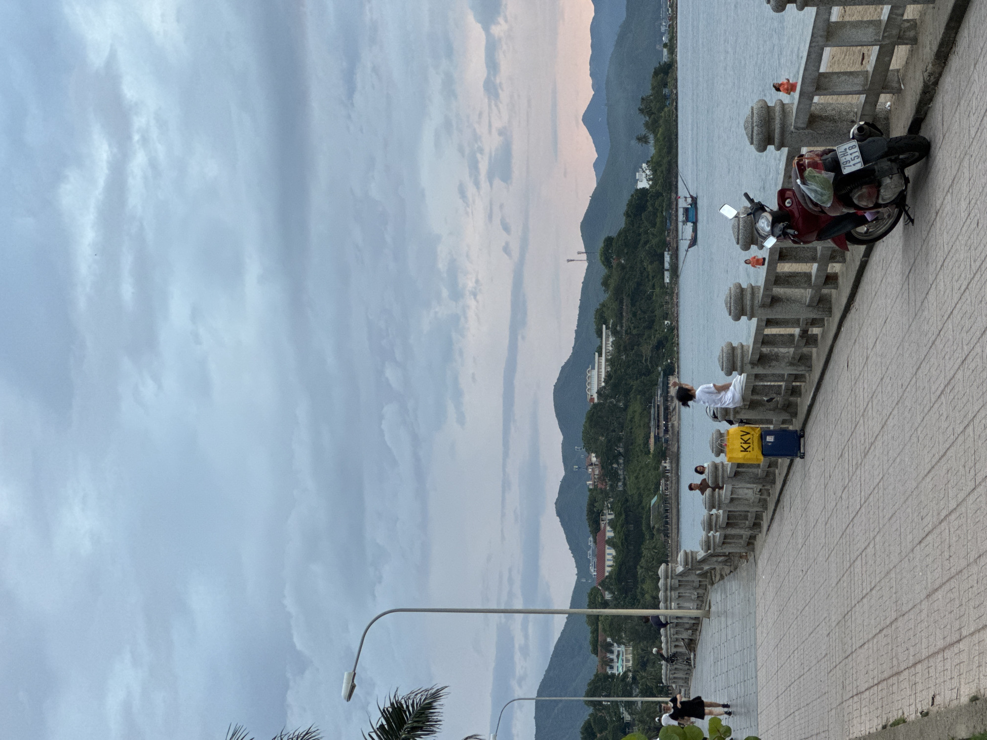 This is a medium-shot, full-frame photograph taken at dusk on a paved promenade in Nha Trang, Vietnam. The promenade runs alongside a body of water, with a shoreline visible on the right. In the background, rolling green hills covered in trees and scattered buildings rise up, with a hazy, overcast sky above.
On the promenade, a woman in a black dress walks towards the left of the frame. Further along, a person sits on a stone railing facing the water, and a bright yellow "KKV" bag is placed on a blue suitcase next to them. A red motorcycle is parked on the promenade in the foreground on the right, with its license plate reading "79-H4 1518". In the water, a small boat with fishing poles extends from its sides is visible, and two people in orange tops are standing in the shallow water near the shoreline. A street lamp arches over the promenade in the middle ground, and palm fronds are visible on the left edge of the frame. The scene conveys a sense of calm and quiet evening activity.