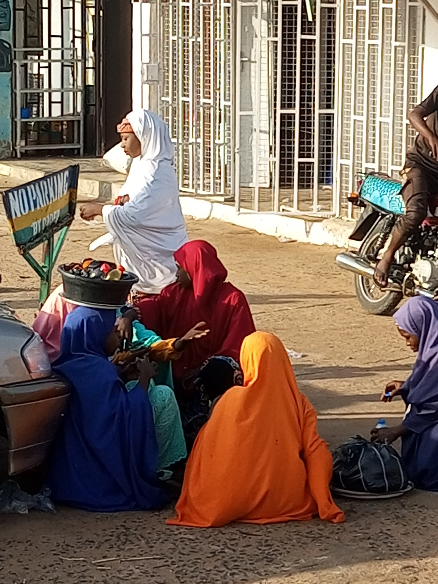 In Gombe, Nigeria, a group of people, primarily women in traditional Muslim attire, are gathered near a street corner. A "NO PARKING" sign is prominently displayed on the left side of the image, indicating a public area. Several individuals are seated on the ground, engaged in various activities. One woman, wearing a white hijab, stands and appears to be attending to a basket filled with items, possibly food or goods for sale. Another woman in a red hijab is seated and gestures with her hands, suggesting she is interacting with others. A black bag and a white bottle are visible near the seated individuals, along with a motorcycle parked in the background. The scene appears to be a typical street market or gathering point, reflecting the local culture and daily life in Gombe.