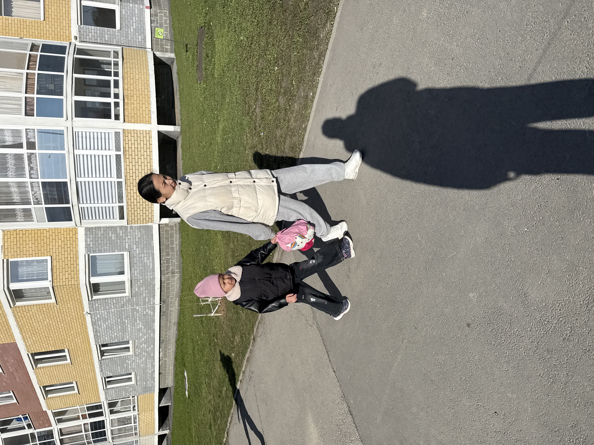 An outdoor scene features two individuals, an adult and a child, standing on a paved surface next to a grassy area. In the background is a multi-story residential building with sections of yellow and grey brickwork, featuring numerous windows and balconies.
The adult, likely a woman, stands on the right, wearing a light beige or off-white puffer vest over a grey long-sleeved top and grey sweatpants, paired with white sneakers. Her dark hair is pulled back, and she is looking downwards.
Standing to the adult's left is a child, likely a girl, wearing a black jacket with a light-colored, possibly faux fur, collar, dark jeans with some embroidered details, and dark sneakers. She has a pink knit beanie on her head and is holding a pink bag adorned with a unicorn design. She appears to be looking towards the adult or the bag.
The activity appears to be a casual standing or posing moment. The bright sunshine indicates it is daytime, with long, distinct shadows suggesting a morning or late afternoon sun. The weather is clear and sunny. A small red circular sign with a white horizontal bar, possibly indicating "No Entry" or "No Stopping," is faintly visible on the building in the background. The architectural style of the building is typical of urban residential areas, consistent with locations such as Yekaterinburg, Russia. Emotions of the subjects are neutral.