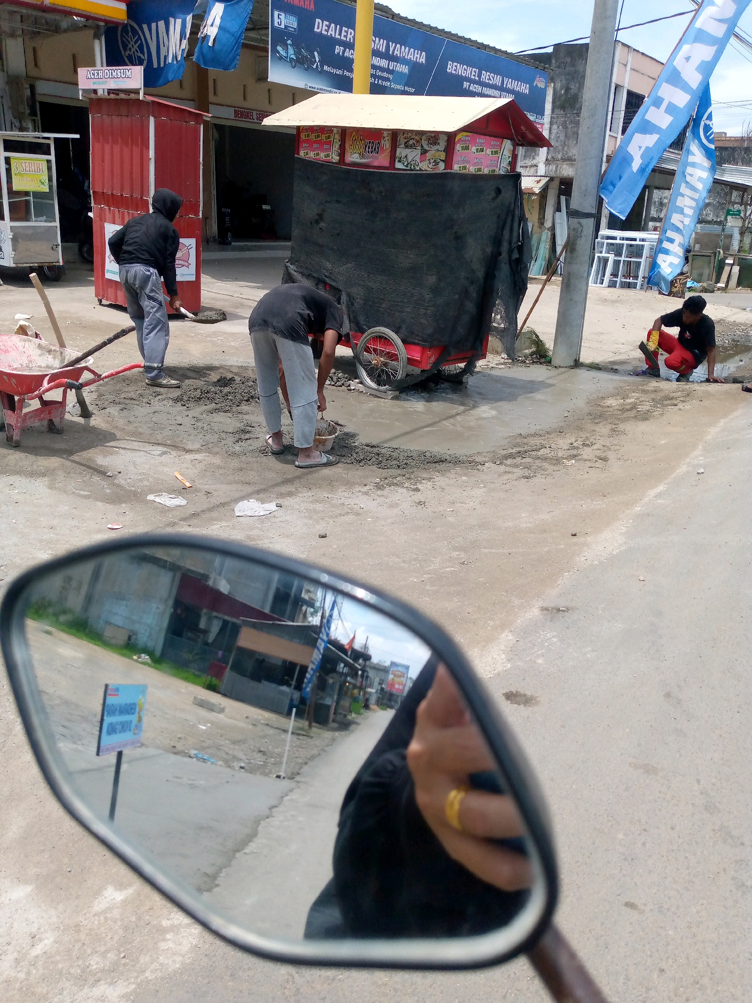 In Blangpeuria, Indonesia, two individuals are engaged in construction work on a dirt road. One person, wearing a black hoodie and grey pants, uses a shovel to move cement. To their right, another individual, in a black shirt and red pants, kneels and works with cement in a small bucket. A red wheelbarrow with construction materials is nearby. In the background, several buildings are visible, including a red structure with "ACER DIMSUM" signage and a Yamaha dealership. Blue Yamaha flags and other banners hang from poles. The scene is partially reflected in a motorcycle's side-view mirror in the foreground, which shows a person wearing a black headscarf with a gold ring on their finger.