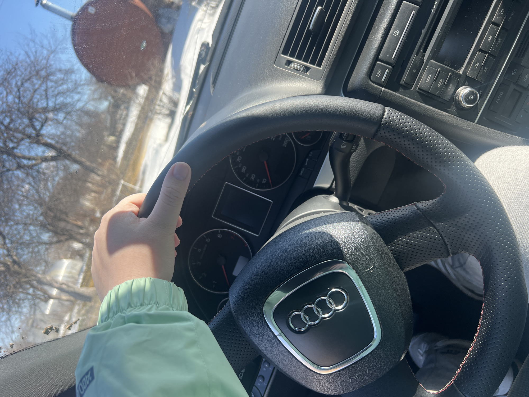 Here is a detailed description of the image:
**Scene:** The image captures the interior of an Audi car from the driver's perspective. The focus is on the steering wheel adorned with the Audi emblem, part of the dashboard, and the driver's hand gripping the steering wheel.
**People:** Only the driver's hand and part of their light-green jacket are visible. No further information about the driver can be ascertained from the image.
**Subjects & Objects:**
* Audi Steering Wheel: Black leather with red stitching and a central Audi emblem.
* Dashboard: Features air vents and an infotainment system with buttons and a CD slot.
* Instrument Cluster: A partially visible speedometer and other gauges.
* Exterior: A glimpse of a blurred outdoor scene can be seen through the windshield, including trees and buildings.
**Actions & Interactions:** The driver is holding the steering wheel, indicating they are likely driving or about to drive.
**Environment:** The exterior scene suggests a potentially residential or suburban area, possibly in winter due to the bare trees.
**Location:** It is impossible to confirm if the image was taken in Chaikovsky, Russia as the image does not provide conclusive evidence. The outdoor background is too blurred to be used to confirm it.