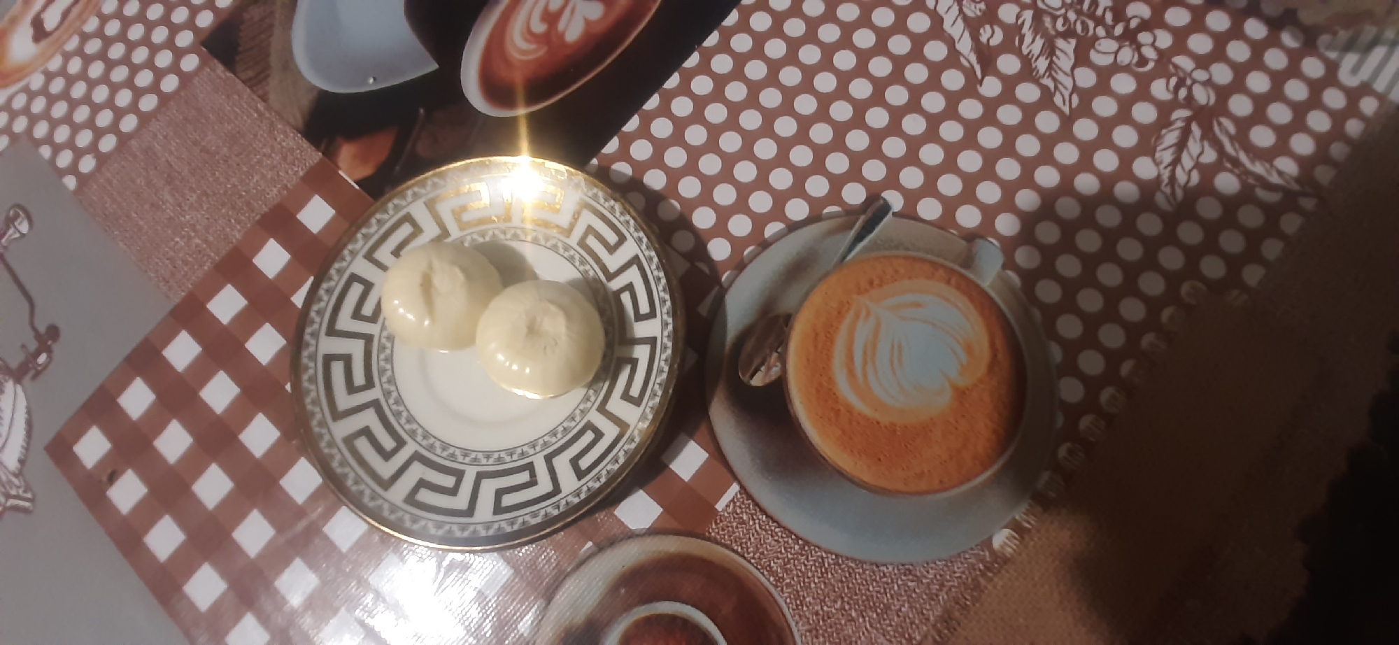 The image shows two small, light-colored truffles on a decorative plate. The plate has a gold rim and a Greek key pattern in black. Beneath the plate is a brown and white checkered placemat. To the right of the plate, there is another placemat with white polka dots on a brown background, and a partial image of a latte art coffee cup. The overall setting appears to be a table. There are no people or specific landmarks visible that would indicate a location in Yessentukskaya, Russia. The image is a close-up shot and the perspective is from above, looking down.