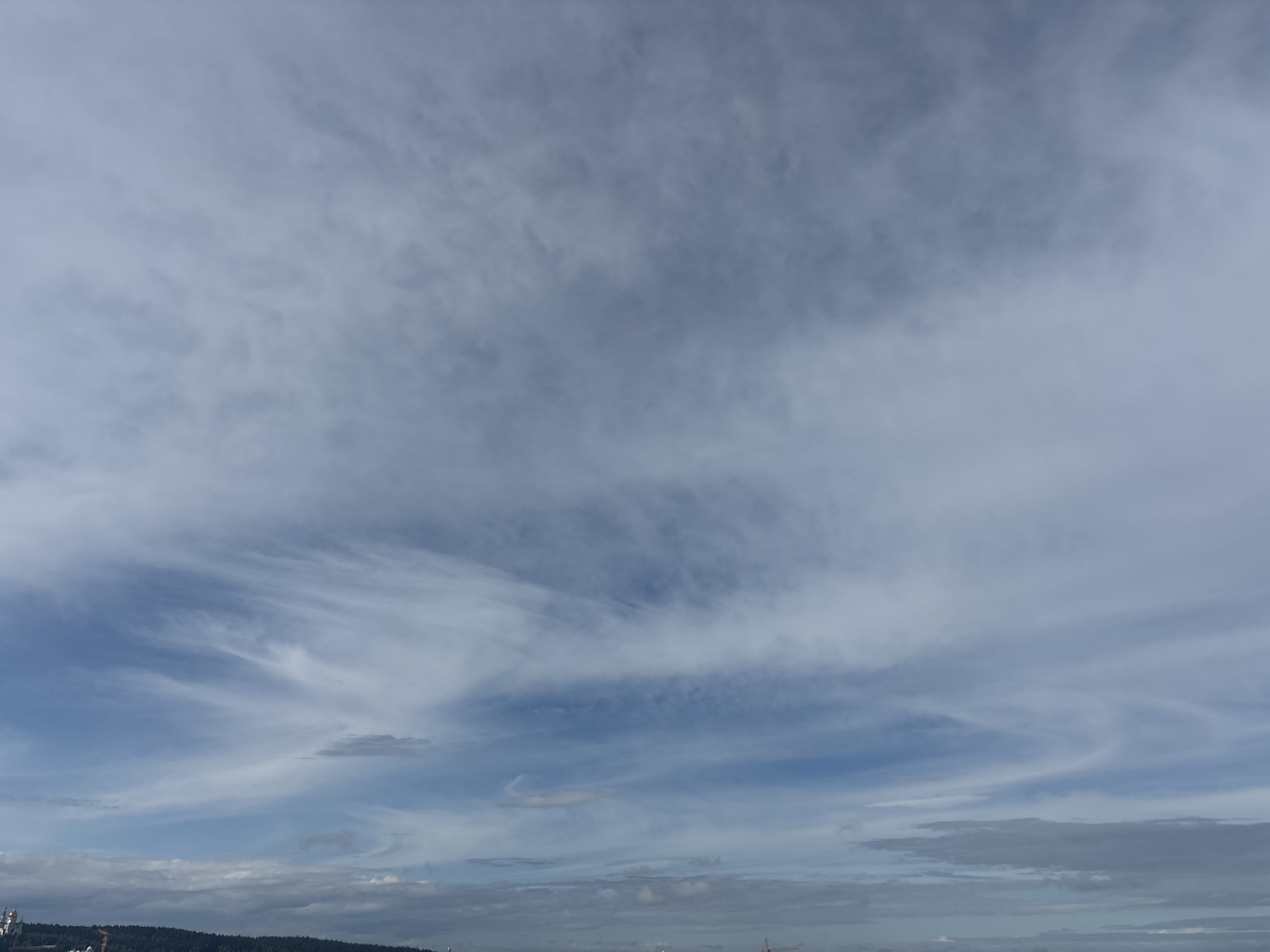The image depicts a wide-angle view of the sky over a landscape. The sky is predominantly filled with clouds, ranging from thin, wispy cirrus clouds in the upper portion to denser, more textured clouds in the lower sections. The clouds exhibit various shades of gray and white, with patches of blue sky visible in between, suggesting an overcast or partly cloudy day.
The lower part of the image shows a distant horizon line, indicating the presence of land. A dark, tree-covered ridge forms a distinct line against the sky. Along this ridge, several structures are visible, including what appear to be buildings and possibly industrial elements, suggested by taller structures and what might be cranes or other machinery on the far left.
There are no visible people or active interactions in the scene. The environment is characterized by a natural landscape dominated by the sky and a distant treeline with some man-made structures. The overall impression is a quiet, observational view of the sky and the edge of a populated area.
While the image displays a sky and landscape, it does not contain any visual information that would allow for a specific identification of the location as Khanty-Mansiysk, Russia. The elements present are generic to many geographical locations with similar natural and semi-urban or industrial features.
