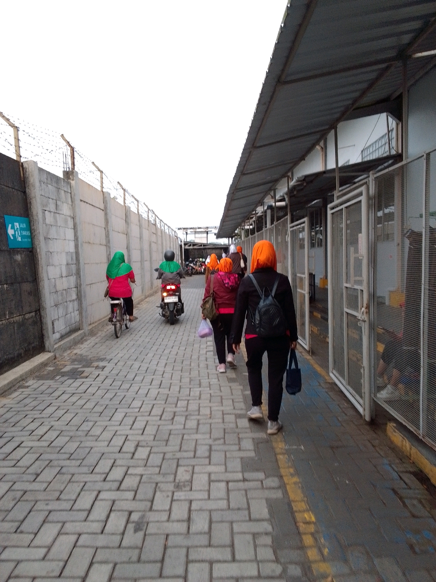 In Sempor, Indonesia, a group of people are walking along a paved walkway next to a grey concrete wall topped with barbed wire. A woman in a green hijab and pink shirt rides a bicycle on the walkway, followed by a person on a scooter wearing a green headscarf and helmet. Further ahead, two women wearing orange headscarves walk in the same direction. The walkway is lined with a building featuring a metal roof and enclosed areas with metal mesh doors. Some of the individuals are carrying bags. The sky is overcast.