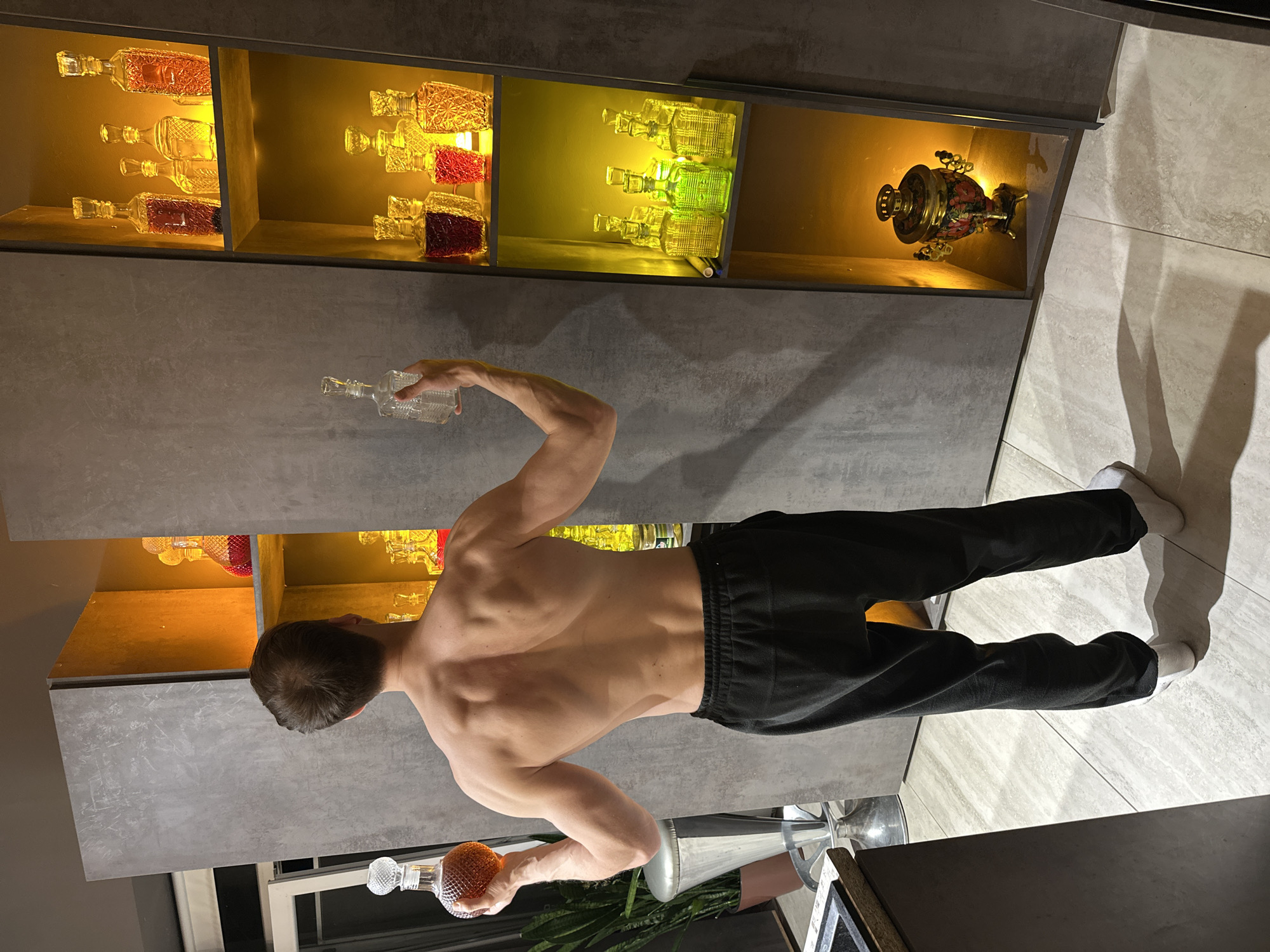 The image shows a shirtless man, viewed from the back, wearing black sweatpants and white socks, standing in a room with a display of illuminated bottles. The man is holding one bottle in each hand. The location is identifiable by the presence of a samovar and distinct liquor bottles suggesting it is in a Russian home, specifically in the city of Kursk. The man is in the foreground, facing towards the wall with the bottle display. The room has tile flooring and a gray cabinet or wall structure.