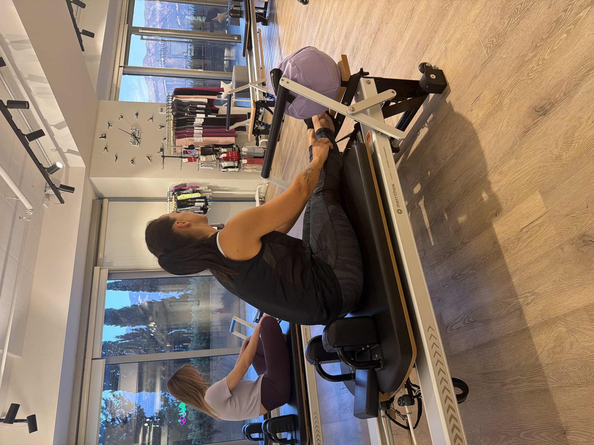 The image shows two women on Pilates reformers in a fitness studio. The studio has large windows offering a view of trees and mountains, likely in Boreti, Montenegro. The woman in the foreground, with her back to the camera, is wearing a black tank top and patterned leggings, performing an exercise on a Merrithew reformer. A lavender-colored Pilates ball rests on the reformer. To her left, another woman with blonde hair, wearing a light purple t-shirt and dark leggings, is also on a reformer. The studio is well-lit with track lighting on the ceiling. Racks of athletic wear are visible in the background, along with a decorative clock on the wall. The floor is made of light-colored wood.