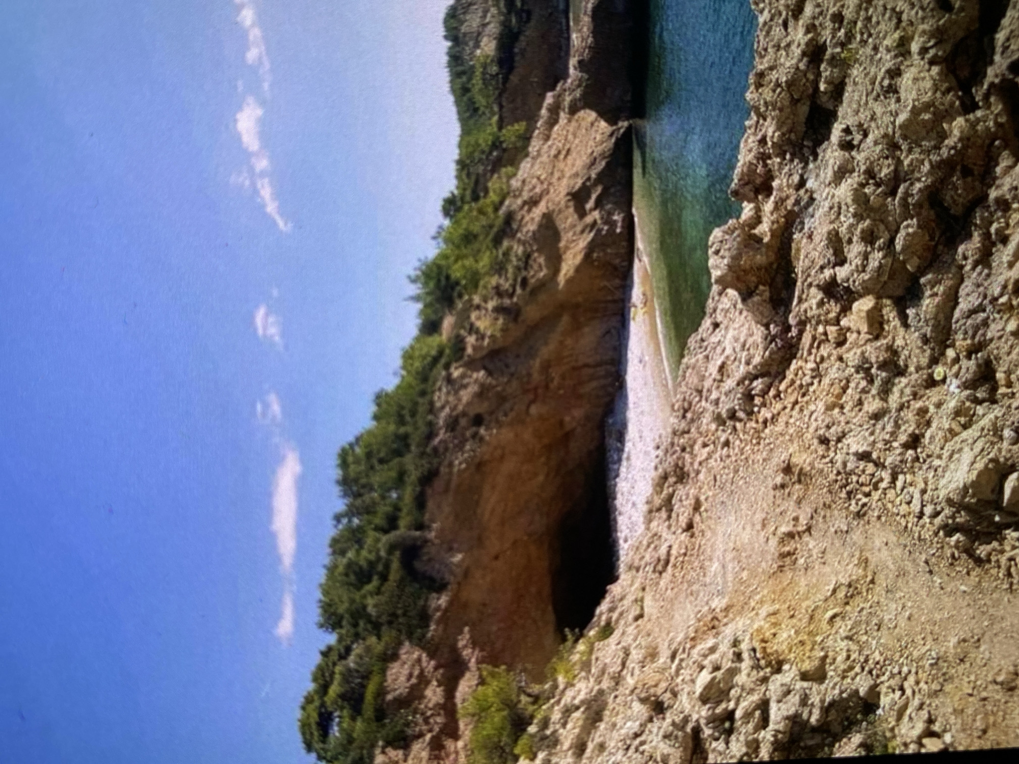 The image presents a natural landscape featuring a rocky coastline or river gorge, bathed in bright sunlight under a clear blue sky with a few wispy white clouds. The photo is tilted, with the horizon running diagonally from upper left to lower right.
On the left side, a towering, rugged cliff face is visible, densely covered with lush green trees and foliage, indicating a vibrant ecosystem. At the base of this cliff, a dark, cavernous opening suggests the presence of a cave or a natural archway leading into the rock formation.
Adjacent to the cliff and extending towards the right, a body of incredibly clear water, shimmering in shades of turquoise and emerald green, signifies a pristine aquatic environment, possibly a calm sea inlet or a wide river. A light-colored, sandy or pebbly shore acts as a narrow transition zone between the water and the base of the cliffs.
The foreground and the right side of the image are dominated by coarse, light-brown to grey rocky terrain, composed of numerous angular stones and rough ground. This rugged slope descends towards the water.
No people or man-made structures are visible. The scene captures a moment of natural serenity and beauty, typical of a bright, sunny day. The clear sky and strong illumination suggest it is midday. This natural setting is located in or near Bankya, Bulgaria.