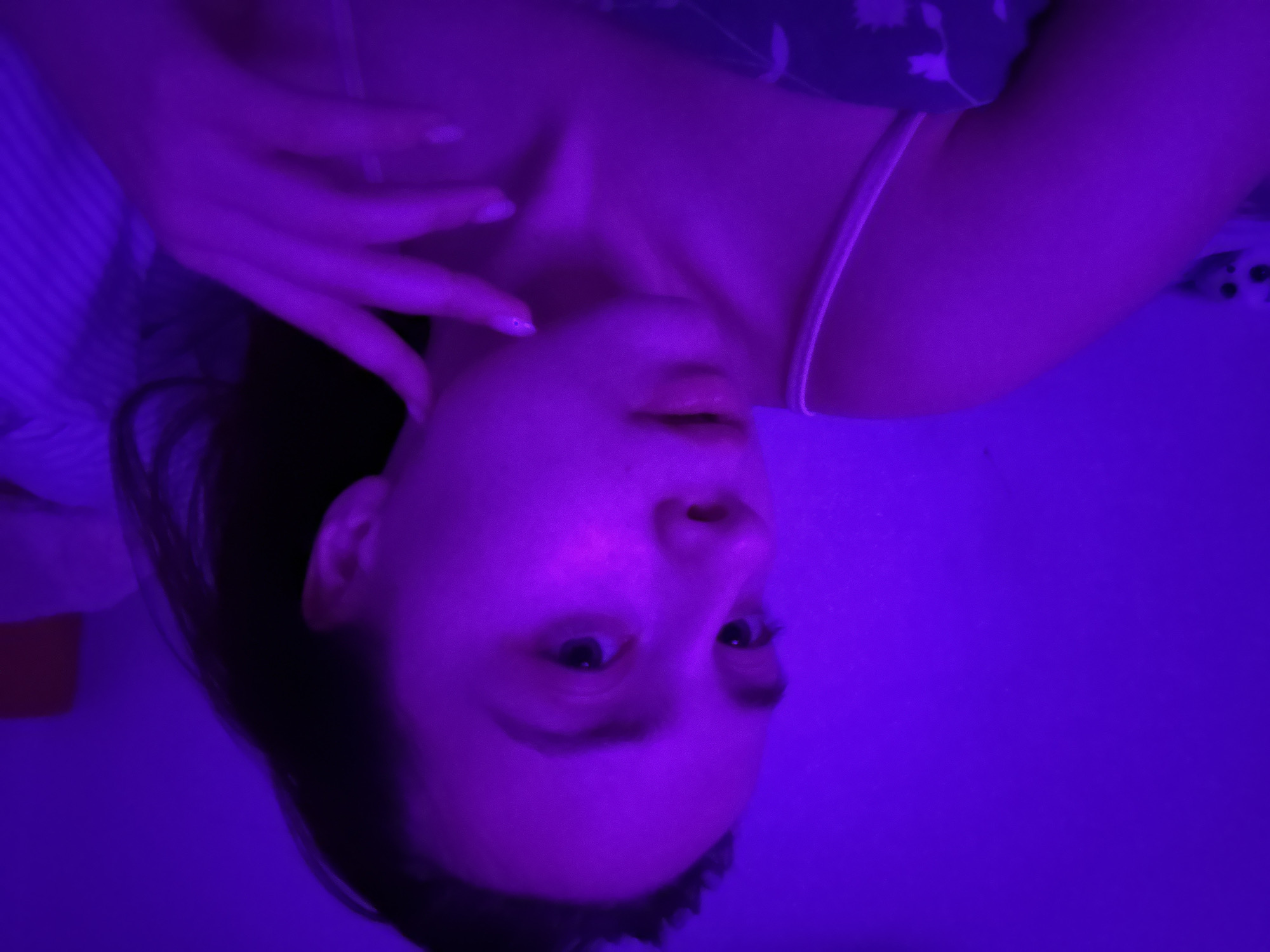 A woman is lying down, possibly on a bed, in a room lit with a strong purple light. She is wearing a camisole top with thin straps. Her face is turned upwards, and she has one hand raised near her neck. The background includes what appears to be bedding with a floral pattern. There is not enough information in the image to determine the location in the city of Прикамский, Russia.
