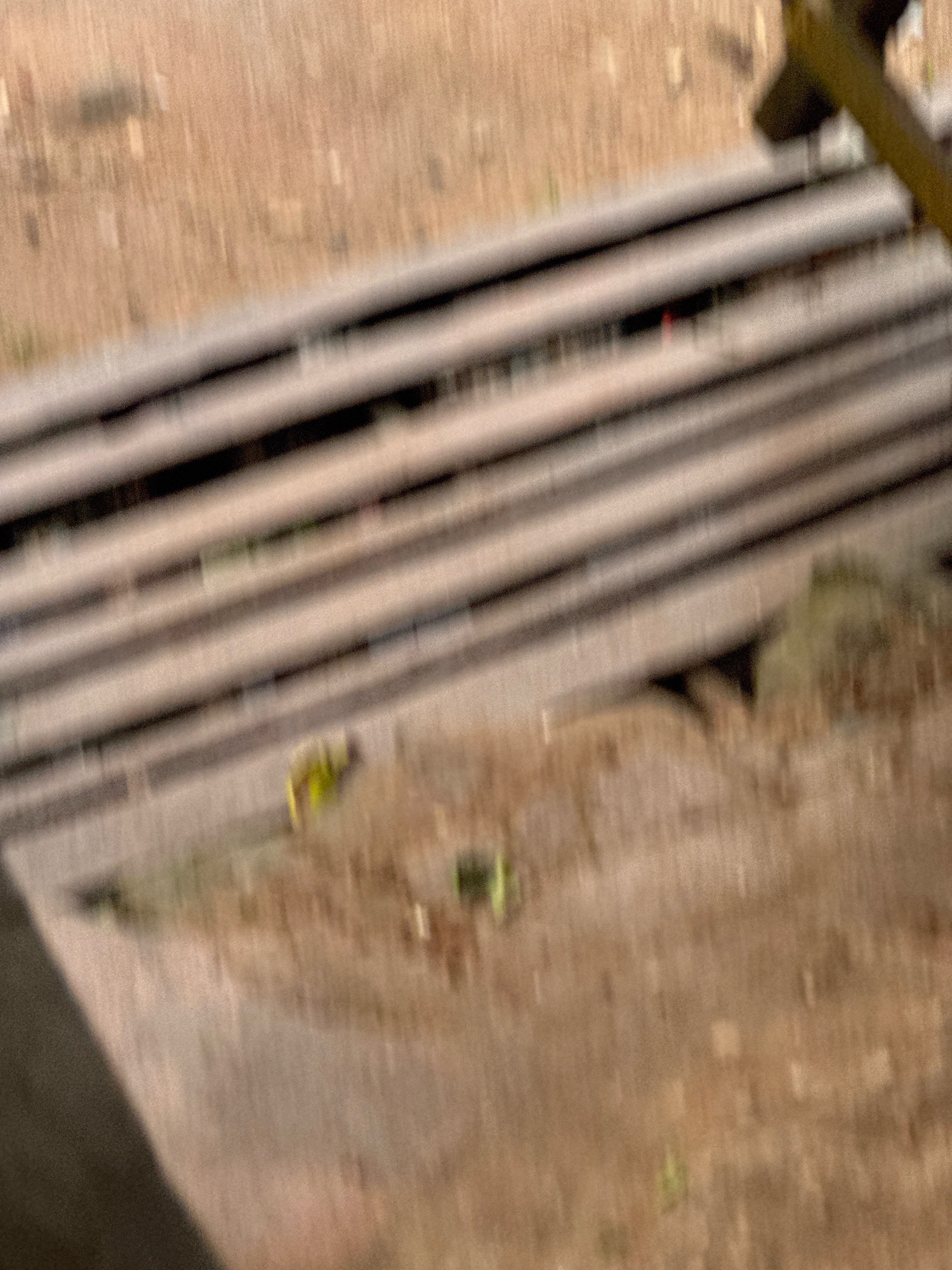 The image, taken from an elevated perspective, shows a section of what appears to be a railway track in Ihiagwa, Nigeria. Multiple parallel metallic rails are visible, running horizontally across the frame. The ground surrounding the tracks is a dusty, sandy terrain with scattered patches of dry vegetation. In the lower portion of the image, near the tracks, there are some blurred green objects, possibly small plants or debris. A small, yellow object is also discernible on the tracks, its shape and purpose unclear due to the image's blurriness. A dark, angular structure occupies the upper right corner of the frame, its purpose and context not fully identifiable. The overall impression is of an outdoor, possibly industrial or transportation-related, environment characterized by its arid conditions and the presence of railway infrastructure.
