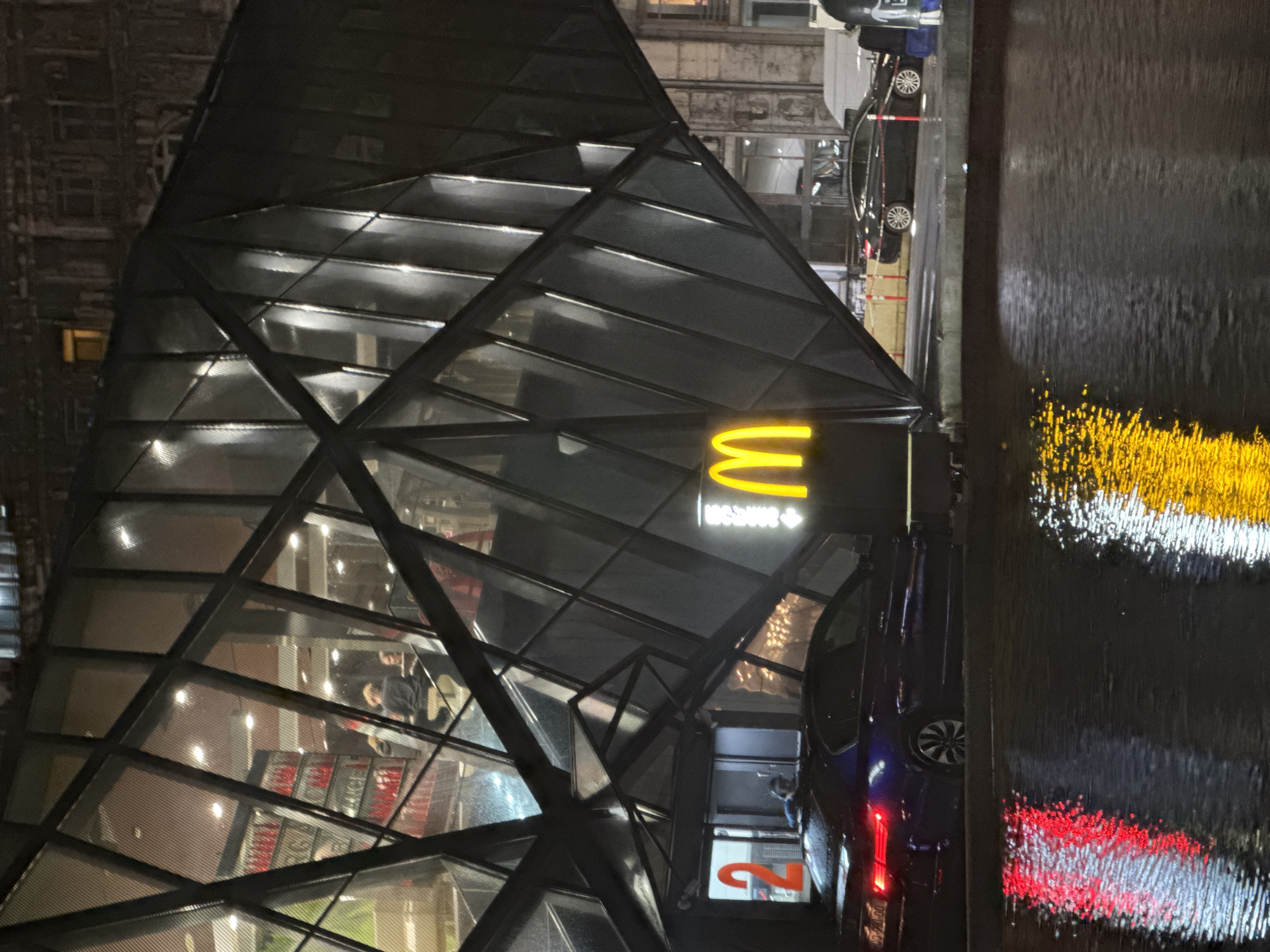Captured at night, the image portrays a McDonald's restaurant situated in an urban setting. The structure of the McDonald's building features a modern, geometric design, largely constructed with glass panels framed by a dark material, possibly metal. The iconic golden arches are illuminated above the entrance.
Inside the restaurant, figures of individuals can be discerned, indicating the presence of patrons.
Adjacent to the McDonald's is a dark-colored car, and further away, another vehicle is also visible on what appears to be a wet street.
To the right of the image, a vertical element is seen, consisting of blurred and refracted lights. This element may be a decorative wall, water feature, or some other kind of artistic installation.
The overall environment appears to be illuminated by streetlights and the lights emanating from the McDonald's itself.