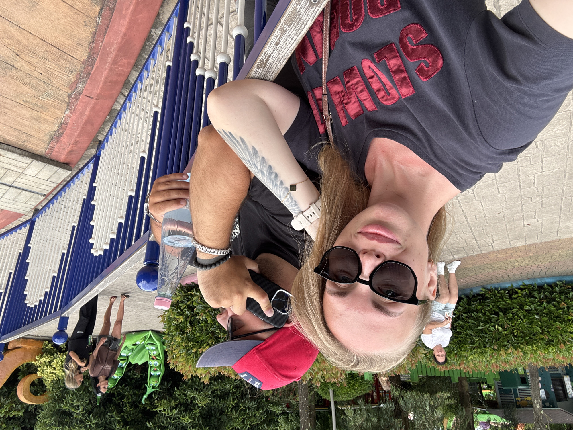 In Sochi, Russia, two individuals, a woman and a man, are captured in a selfie taken from a low angle. The woman, in the foreground, is wearing sunglasses and a dark t-shirt with red lettering. Her left arm, adorned with a tattoo and a watch, rests on a white and blue railing. The man, partially visible behind her, is wearing a red baseball cap and holding a black smartphone to his ear. He is dressed in a dark t-shirt and has a beaded bracelet on his right wrist. In the background, a person in a white t-shirt and shorts is seated on a bench. Further in the distance, two women are walking. The environment appears to be an outdoor recreational area, possibly a park, with lush green foliage and decorative elements like a large green pea pod sculpture. The walkway is paved with stone-like tiles and bordered by wooden planks.