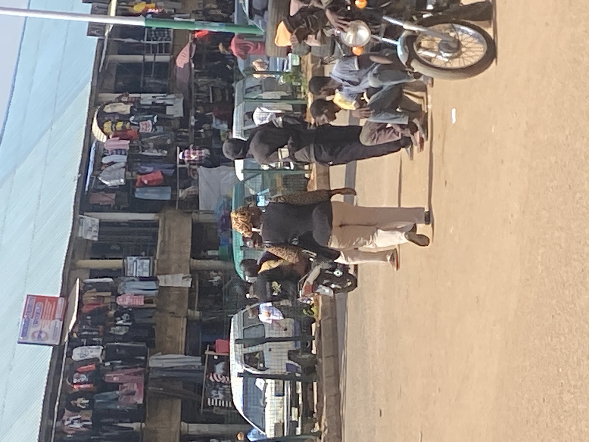 A street scene in Bukuru, Nigeria, shows a marketplace with several people and vehicles. In the foreground, a white van is parked on the left side of the street. To the right of the van, several men are standing and sitting, some near a motorcycle. Further back, a woman is walking away from the camera, wearing a patterned top and light-colored pants. Shops line the street, displaying various clothing items. The street is paved with dirt and sand. The overall impression is of a bustling, everyday market environment in Nigeria.