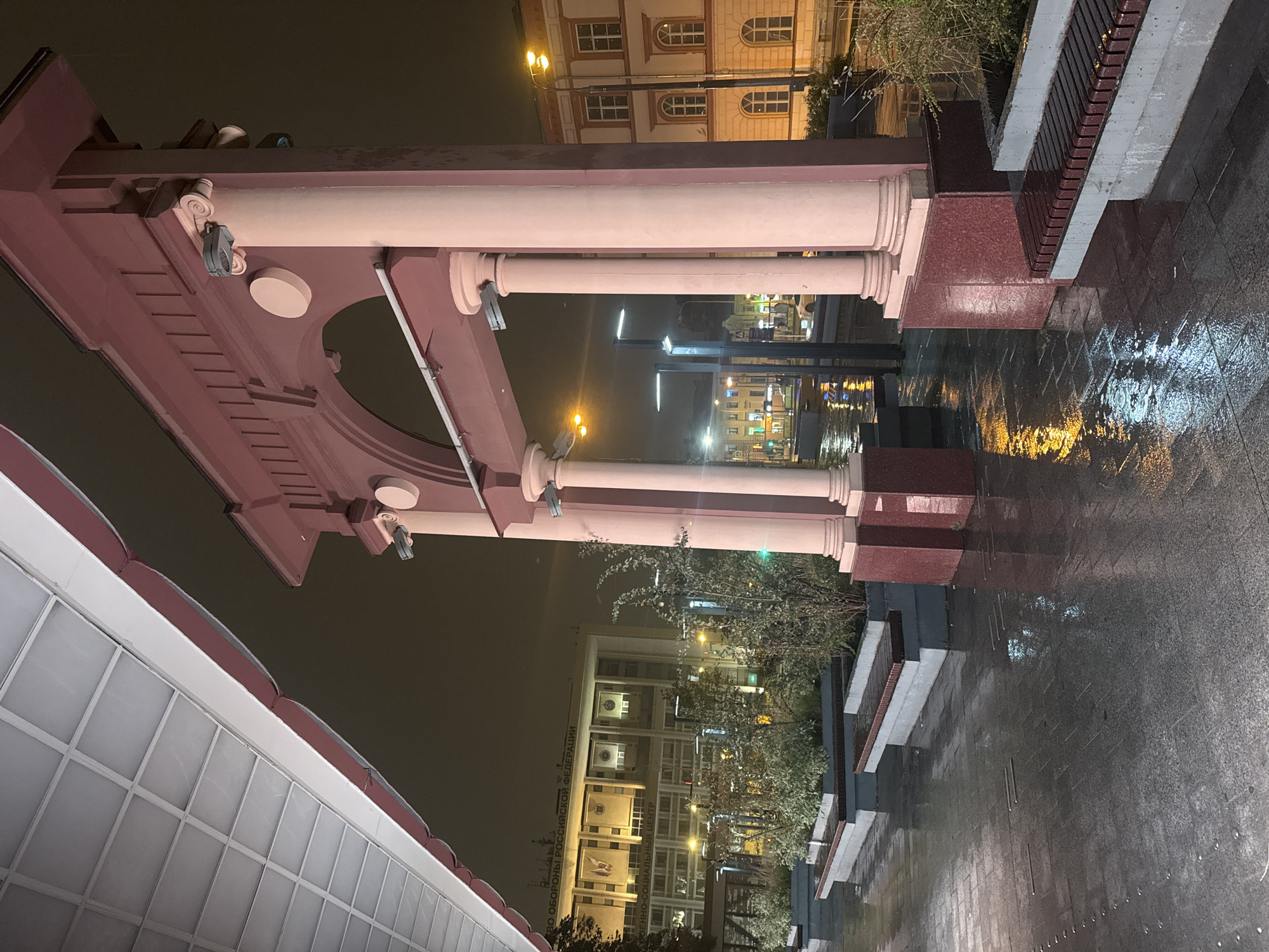 A night scene on a wet, paved walkway in Moscow, Russia, features a decorative archway with large columns. To the left, a building with multiple windows and signage that reads "МИНИСТЕРСТВО ОБОРОНЫ РОССИЙСКОЙ ФЕДЕРАЦИИ" (Ministry of Defense of the Russian Federation) and "ВОЕННО-СОЦИАЛЬНЫЙ ЦЕНТР" (Military Social Center) is visible. The walkway is lined with planters containing bushes and benches. Streetlights cast reflections on the wet pavement. The overall atmosphere is subdued due to the nighttime setting and the rain.